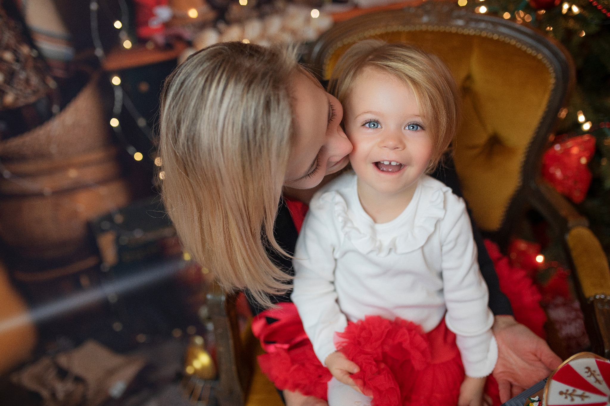 Mama całuje uśmiechniętą córeczkę w czerwonej spódniczce na ozdobnym, żółtym fotelu, z choinką w tle.