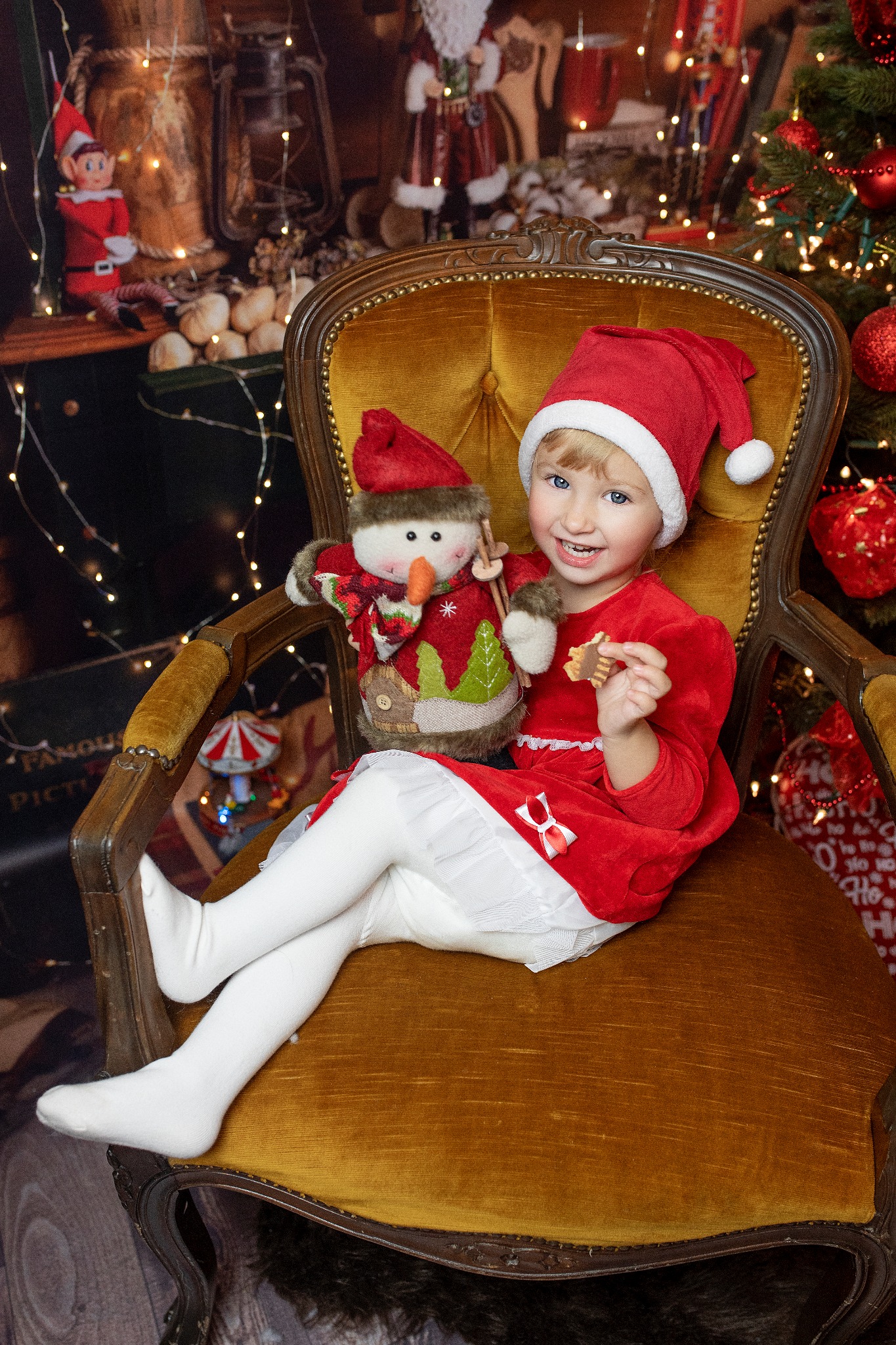 Uśmiechnięta dziewczynka w czerwonym stroju Mikołaja siedzi na złotym, zdobionym fotelu, trzymając pluszowego bałwanka i ciasteczko, na tle świątecznej dekoracji z choinką i lampkami.
