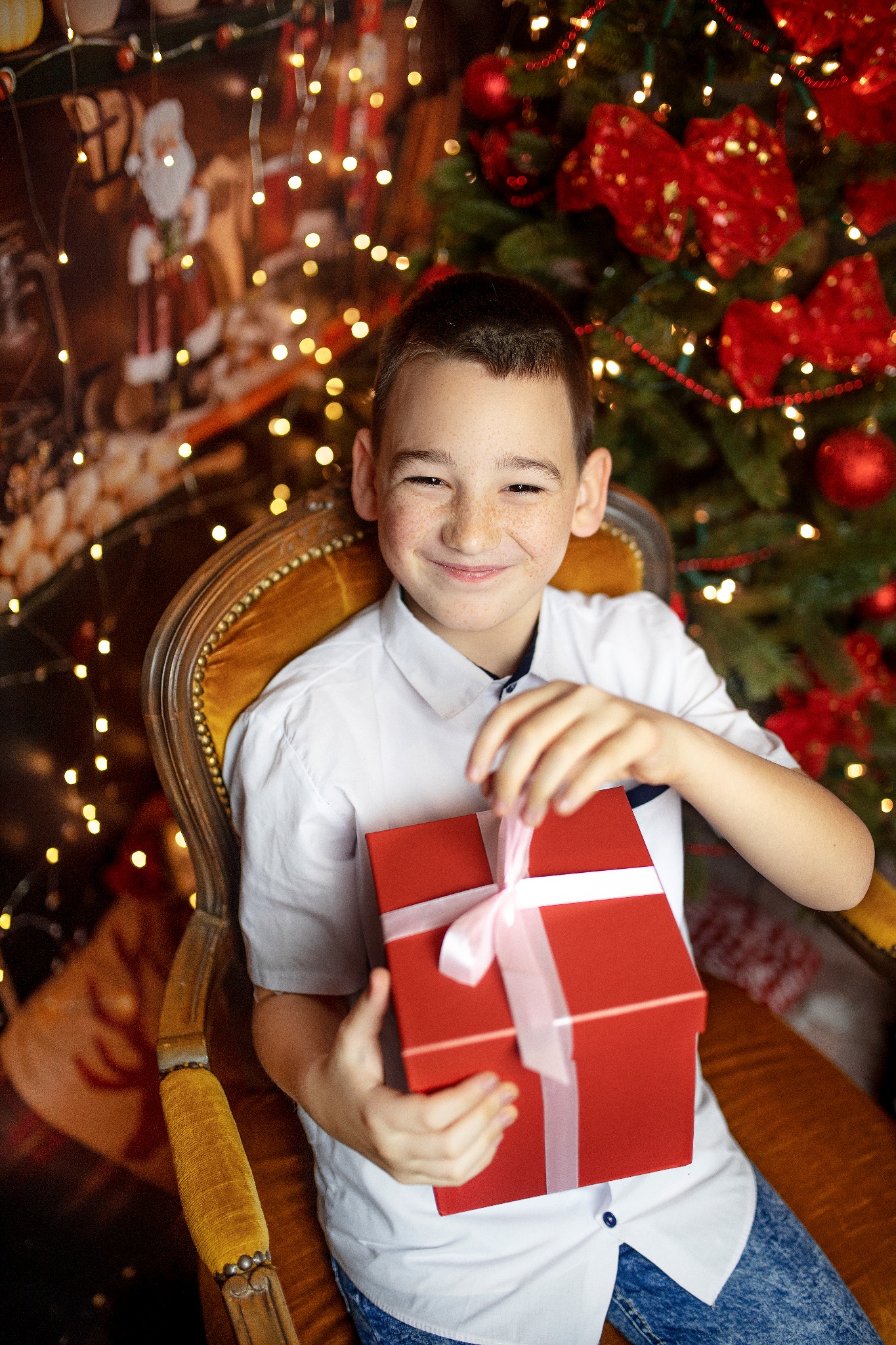 Uśmiechnięty chłopiec z piegami siedzi na fotelu w świątecznej scenerii, trzymając czerwone pudełko z białą wstążką, w tle choinka z czerwonymi ozdobami i lampkami.