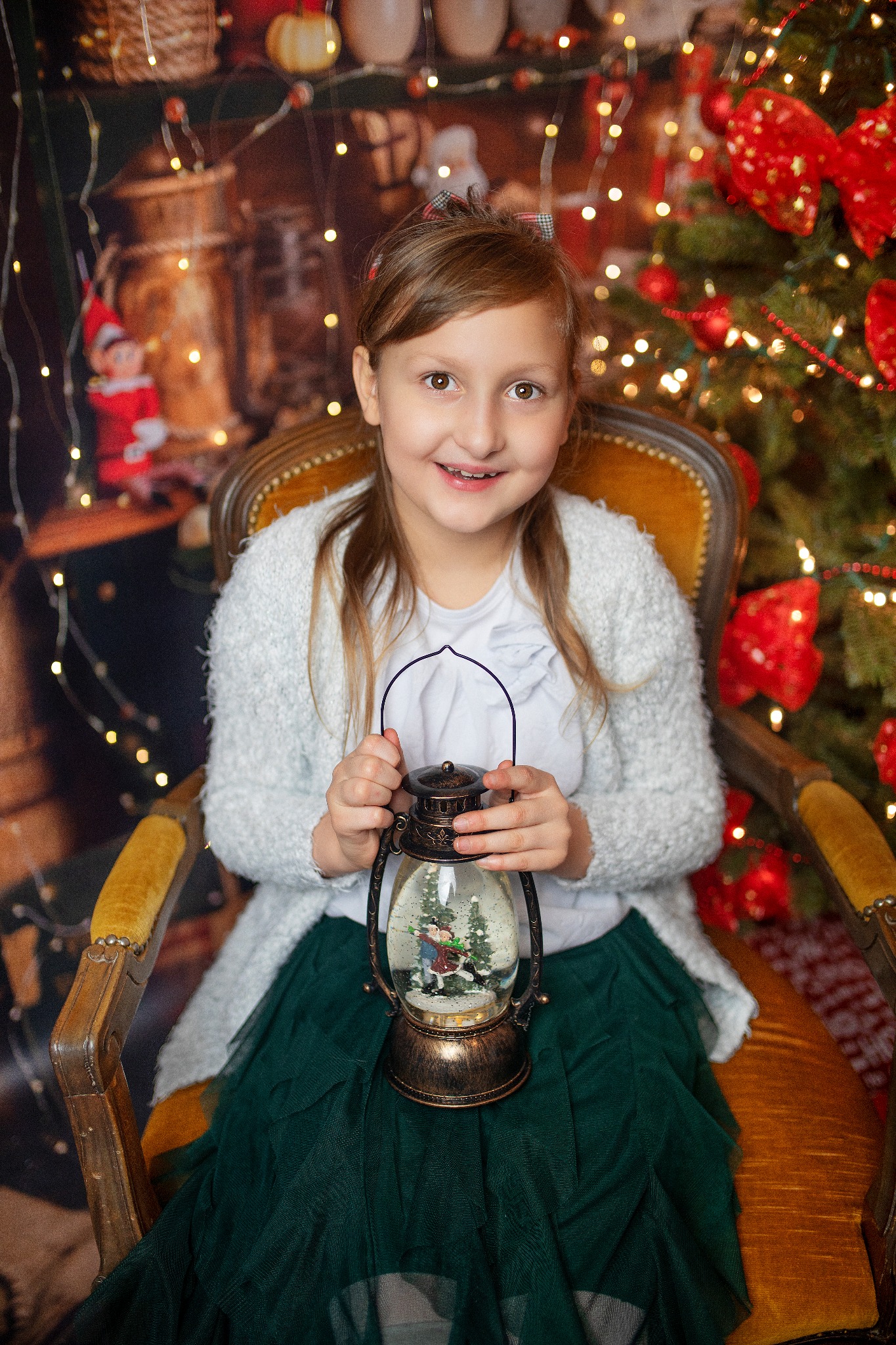 Uśmiechnięta dziewczynka siedzi na fotelu, trzymając lampion ze śnieżną scenką. W tle choinka z czerwonymi ozdobami i dekoracje świąteczne.