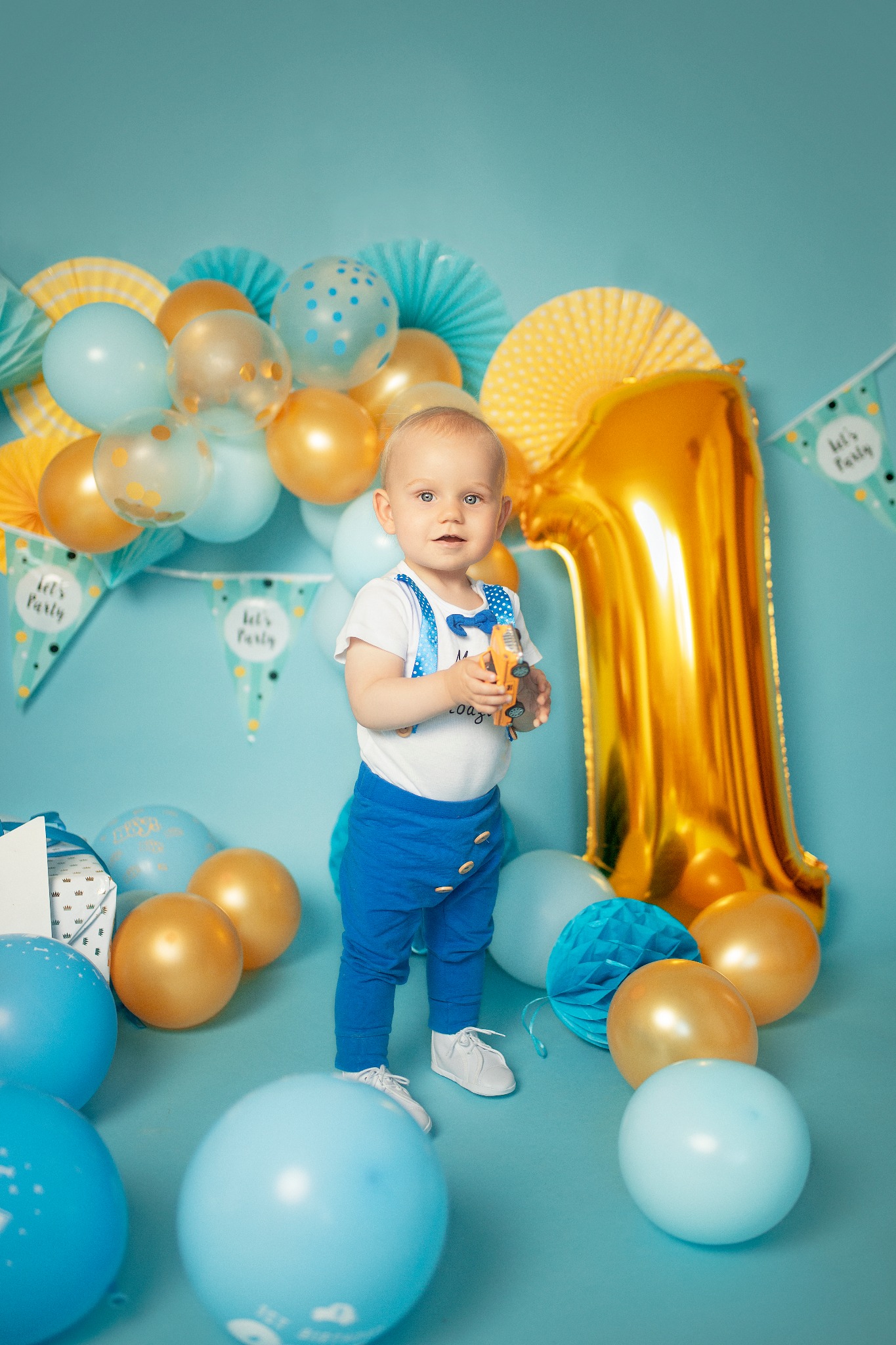 Roczne dziecko w niebieskim stroju z muchą, trzymające zabawkę-autko, pozuje na tle niebieskiego studia z balonami i dekoracjami urodzinowymi, w tym dużą złotą cyfrą '1'.