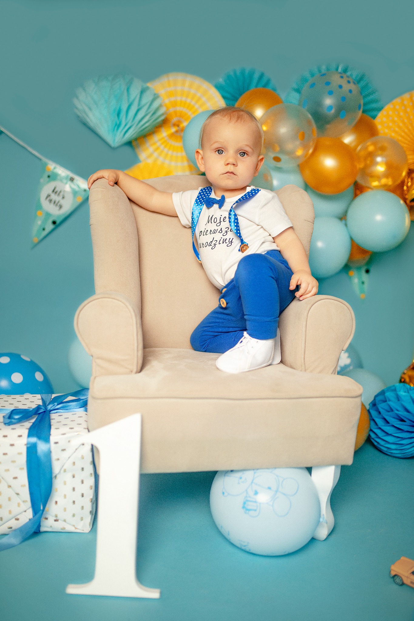 Chłopiec w niebieskich spodniach i białej koszulce z napisem 'Moje pierwsze urodziny' siedzi w beżowym fotelu na tle błękitnych dekoracji urodzinowych, balonów i prezentu z niebieską wstążką, obok...
