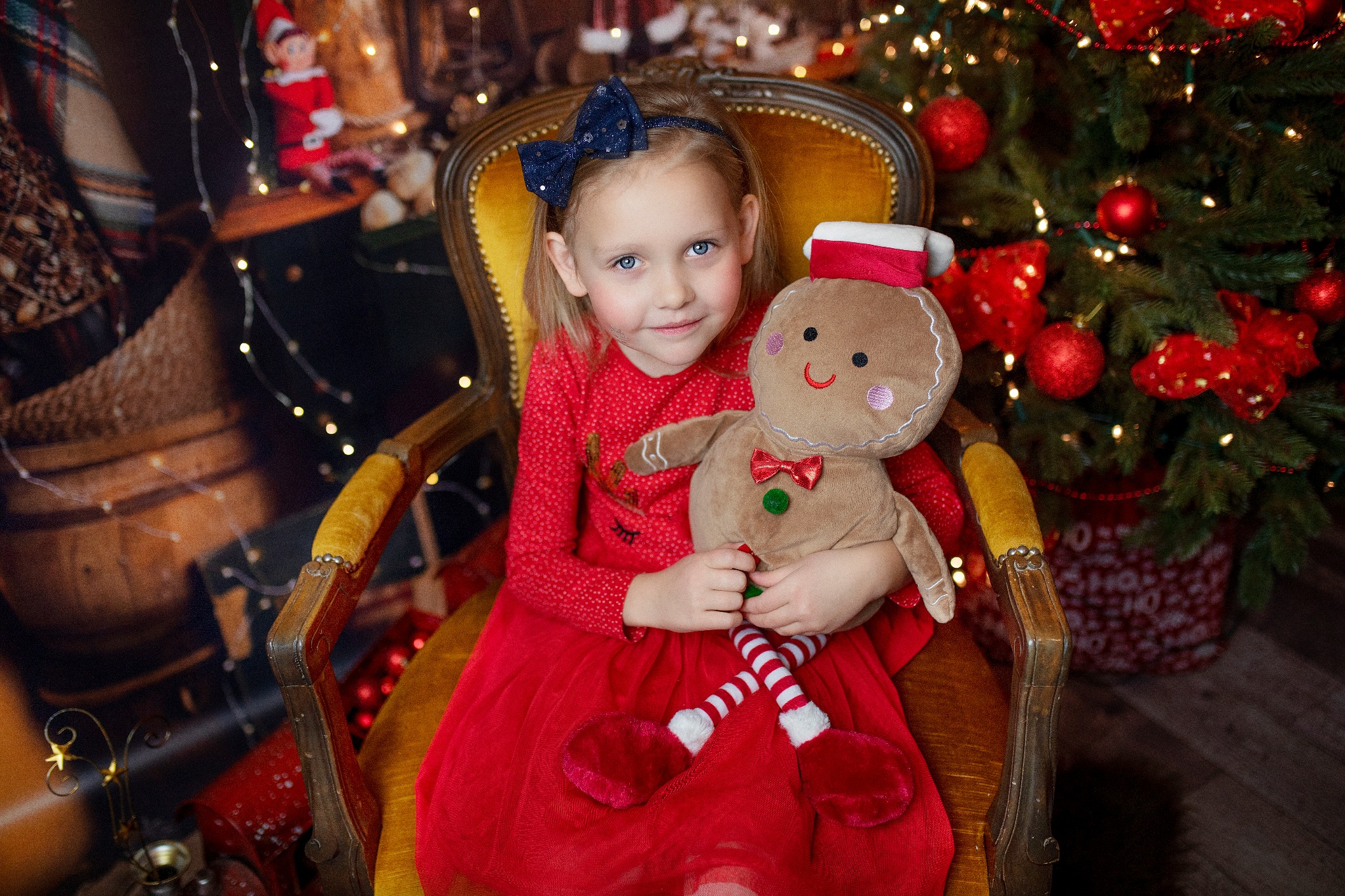Uśmiechnięta dziewczynka z niebieskimi oczami w czerwonej sukience siedzi w fotelu i trzyma pluszowego pierniczka na tle choinki i świątecznych dekoracji.