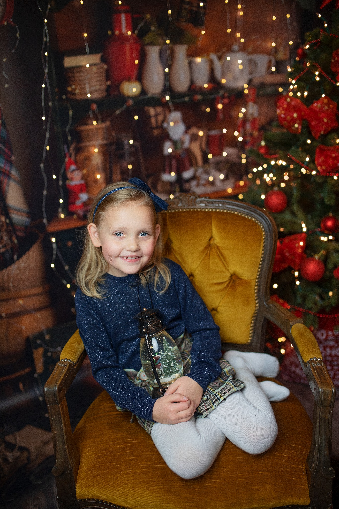 Uśmiechnięta dziewczynka z blond włosami, siedząca na złocistym fotelu w świątecznej scenerii, trzymająca lampion ze śnieżną kulą w środku, w tle choinka z czerwonymi ozdobami i półki z dekoracjami.