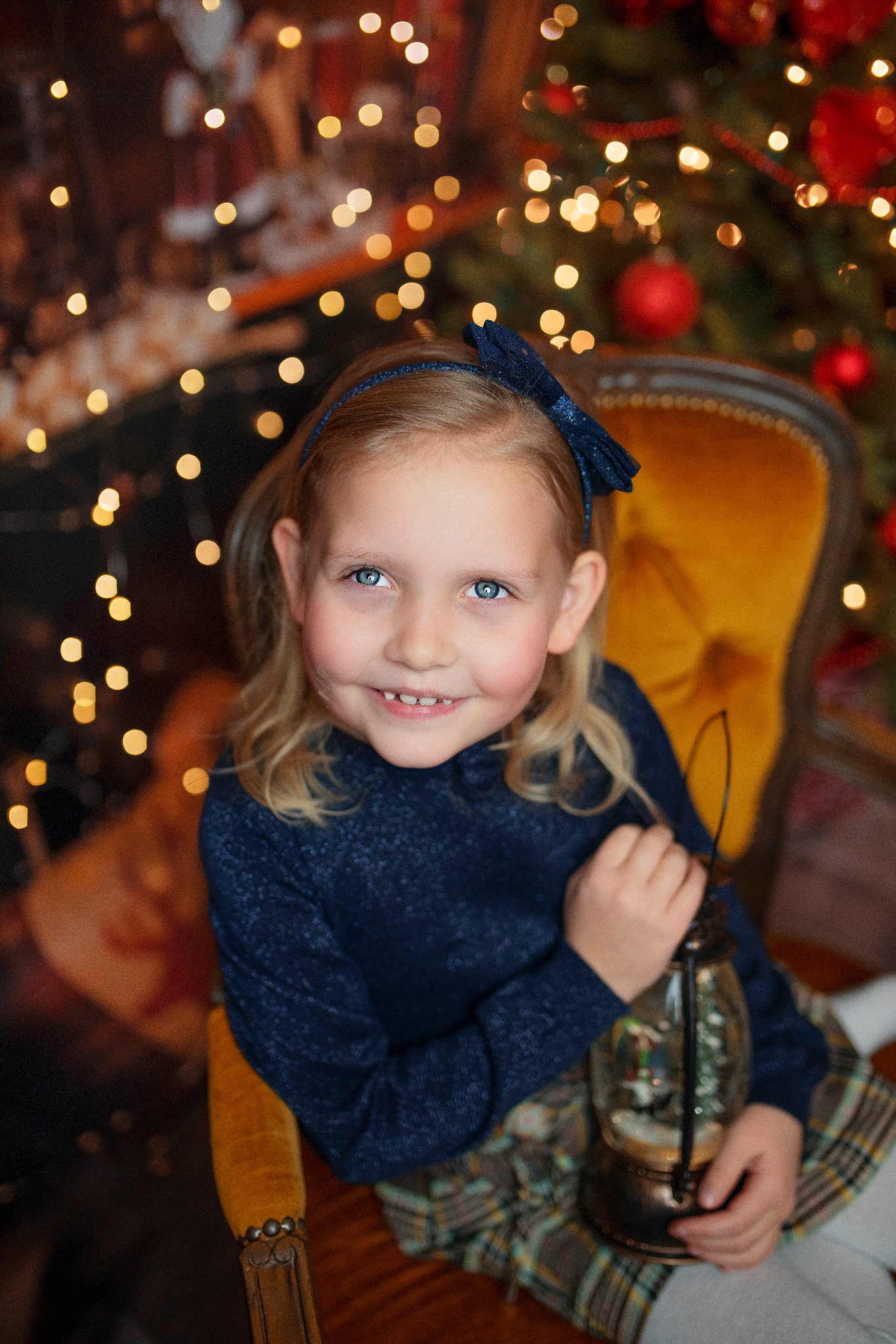 Uśmiechnięta dziewczynka z niebieskimi oczami, siedząca na żółtym fotelu, trzymająca lampion ze śnieżną kulą na tle choinki z lampkami.