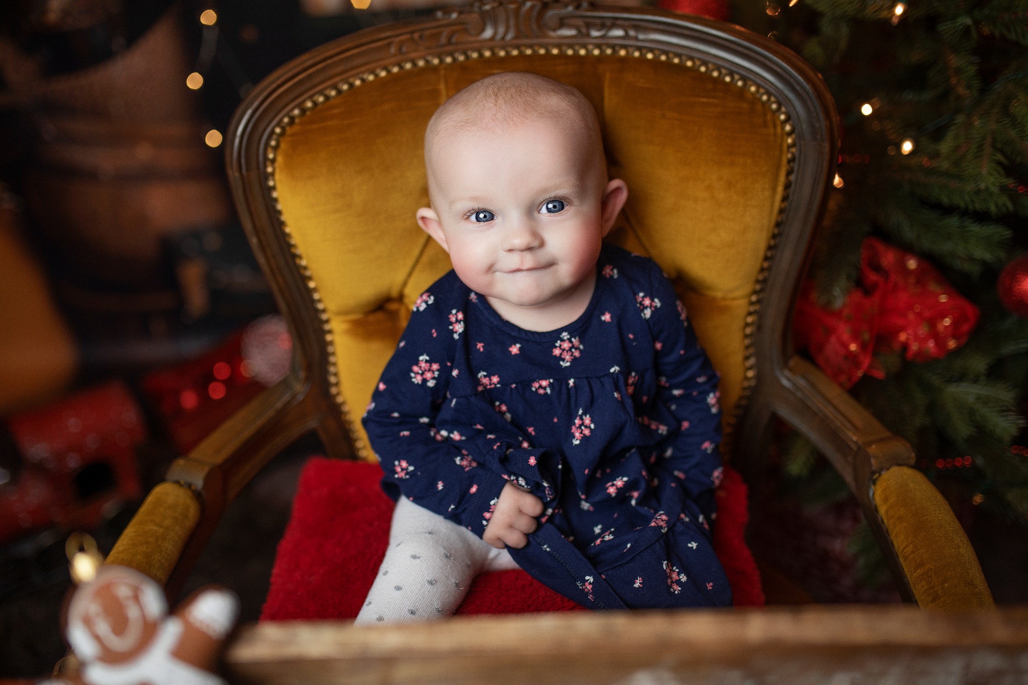 Uśmiechnięta niemowlęca dziewczynka w kwiecistej sukience siedzi w złotym fotelu na czerwonej poduszce, w tle choinka z czerwonymi ozdobami i światełkami.