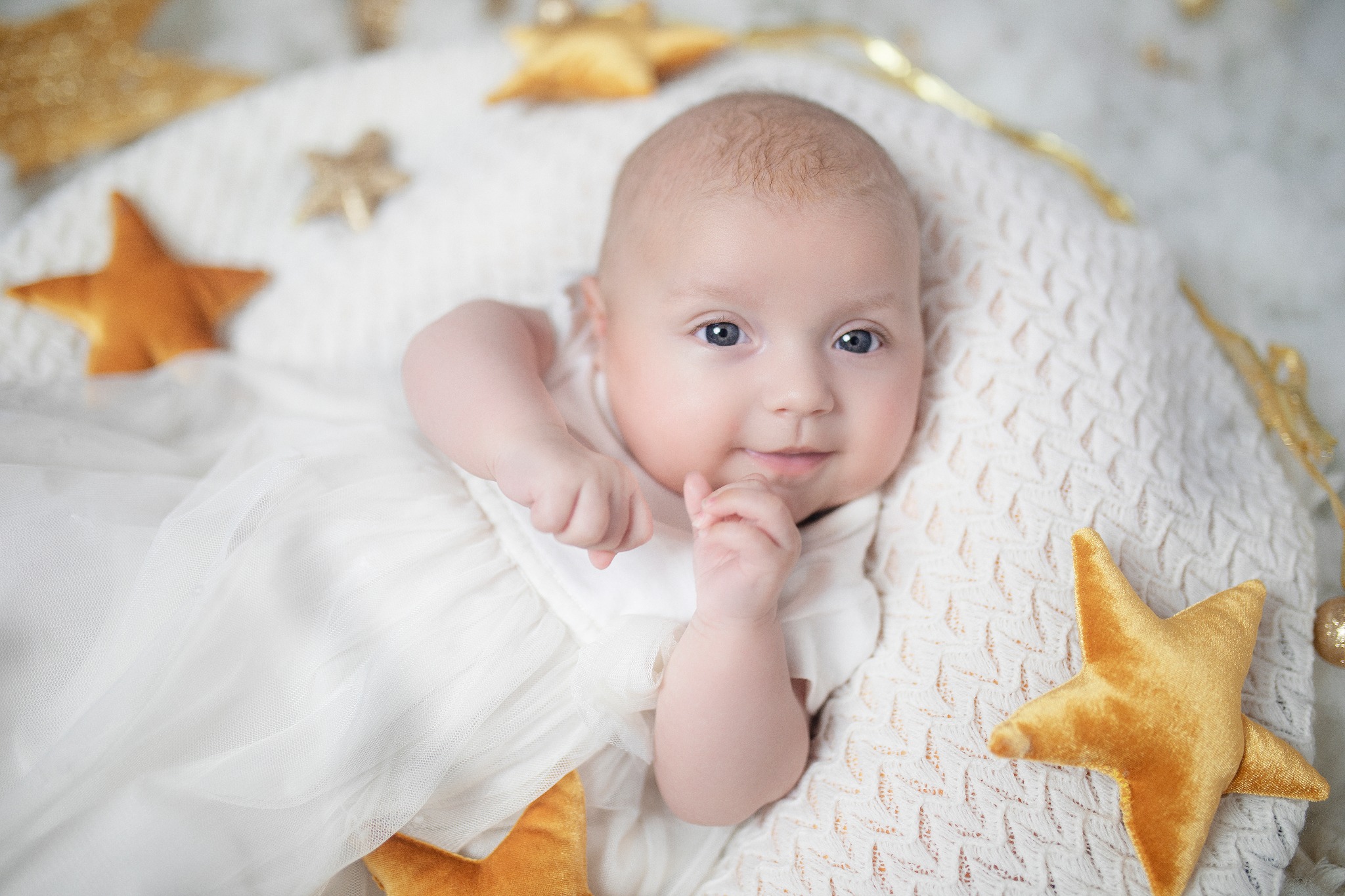 Urocze niemowlę w białej sukience leży na puszystej poduszce w kształcie półksiężyca, otoczone złotymi gwiazdkami, z delikatnym uśmiechem i dużymi niebieskimi oczami.