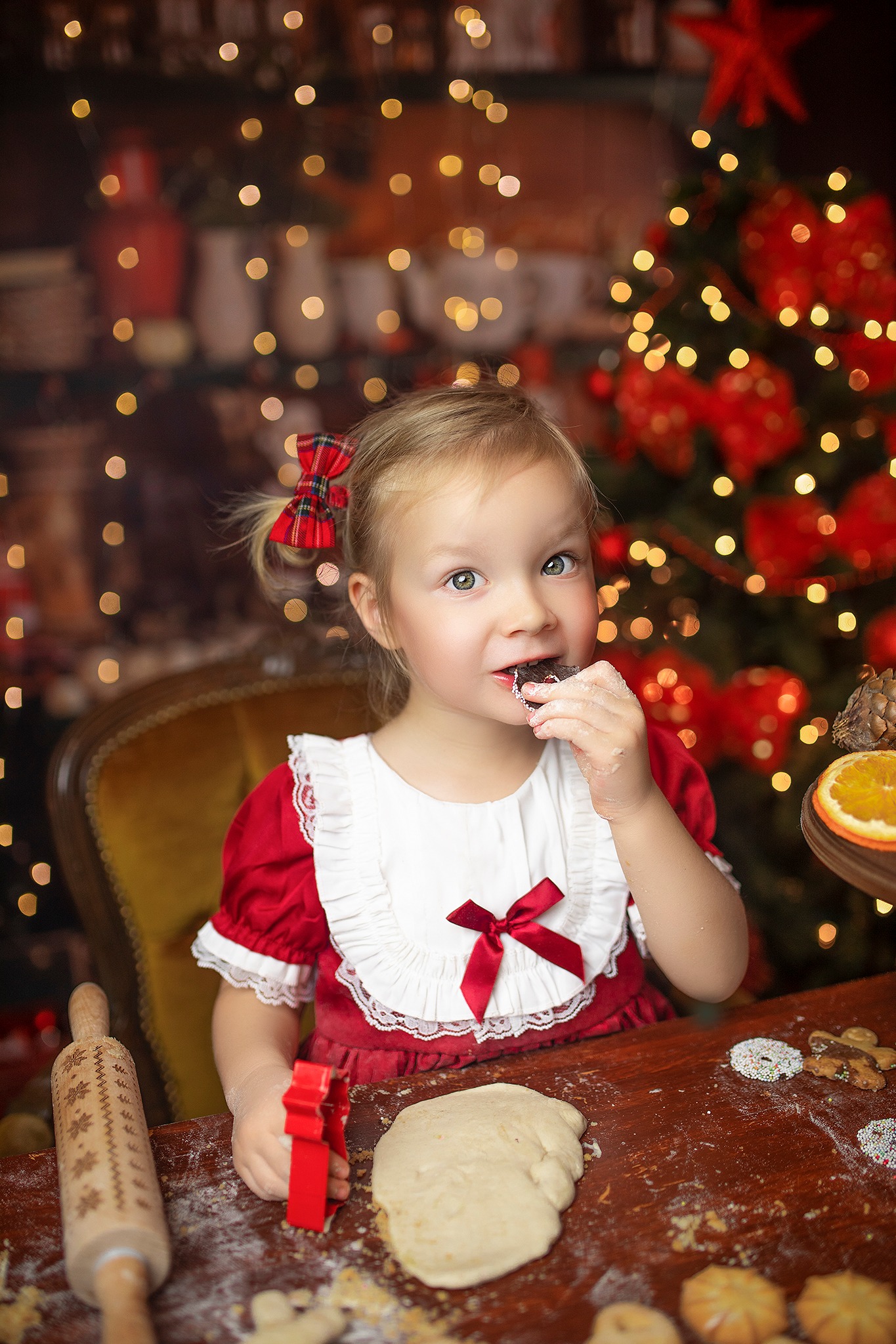 Dziewczynka w czerwonej sukience z kokardą w warkoczu je ciasteczko na tle choinki, obok wałek z wygrawerowanymi wzorami i wykrawacz do ciastek w kształcie gwiazdy.