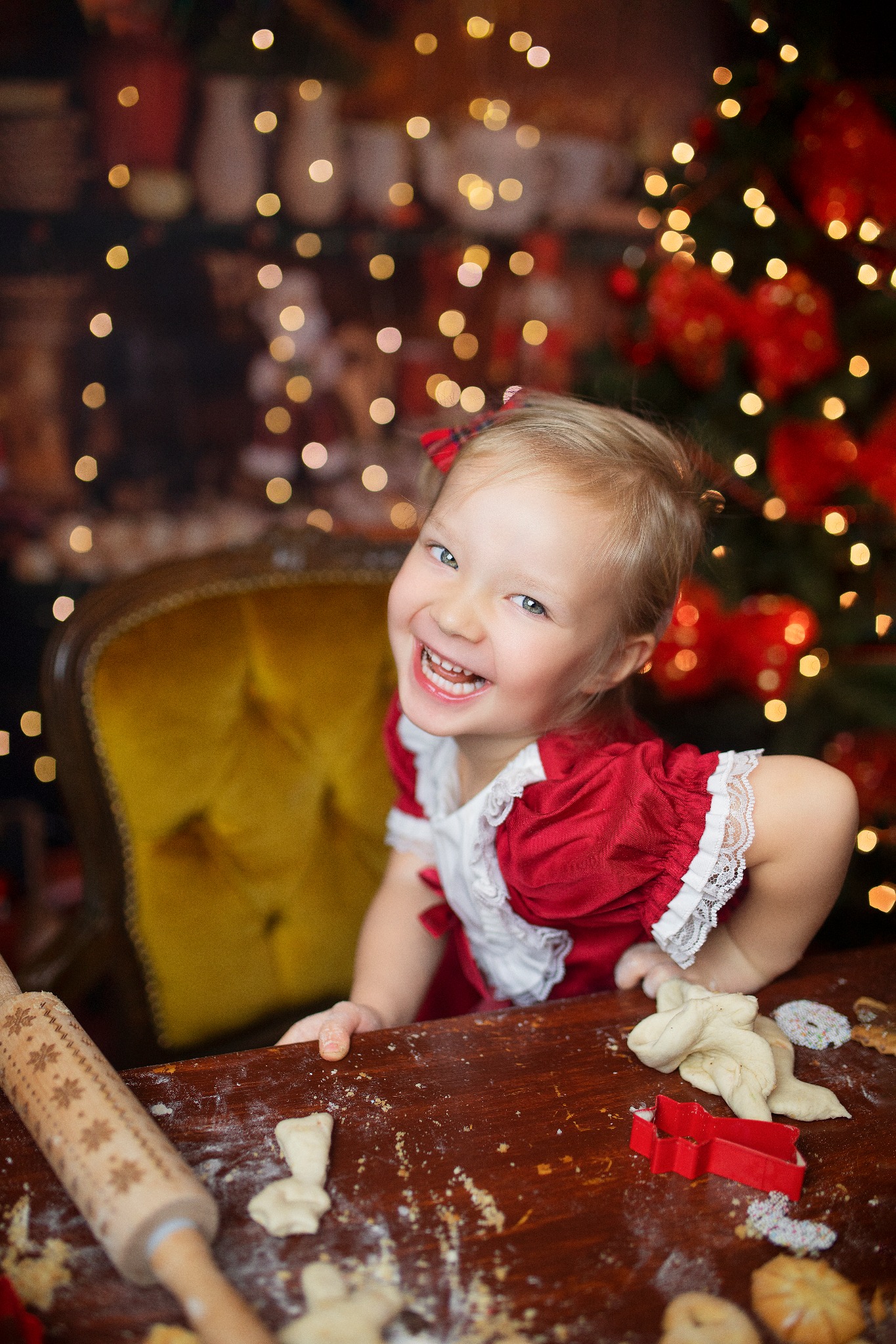 Uśmiechnięta dziewczynka w czerwonej sukience z koronką, siedząca przy drewnianym stole z wałkiem do ciasta i foremkami, w tle choinka z lampkami.