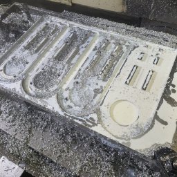 ARMET ADAM GÓRCZYŃSKI - Biały detal z wyfrezowanymi otworami, pokryty wiórami metalowymi, w trakcie obróbki CNC. Widoczna głowica frezarki.