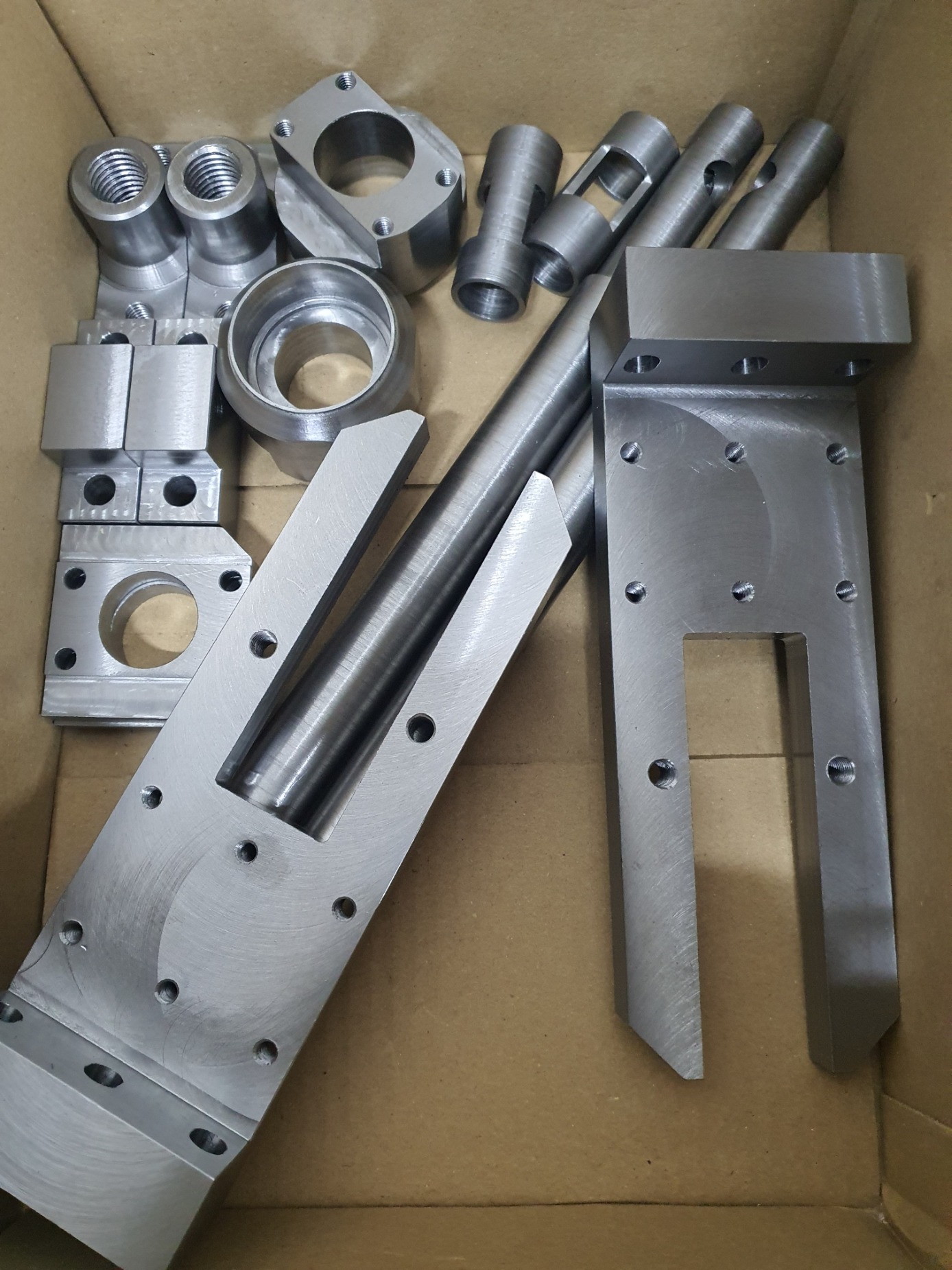 Metalowe elementy po obróbce w kartonowym pudełku. Widoczne gwintowane tuleje, pierścienie, pręty i płyty z otworami. Szlifowana powierzchnia, precyzyjne wykonanie.