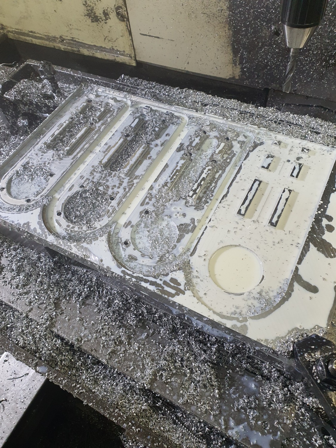 Biały detal z wyfrezowanymi otworami, pokryty wiórami metalowymi, w trakcie obróbki CNC. Widoczna głowica frezarki.