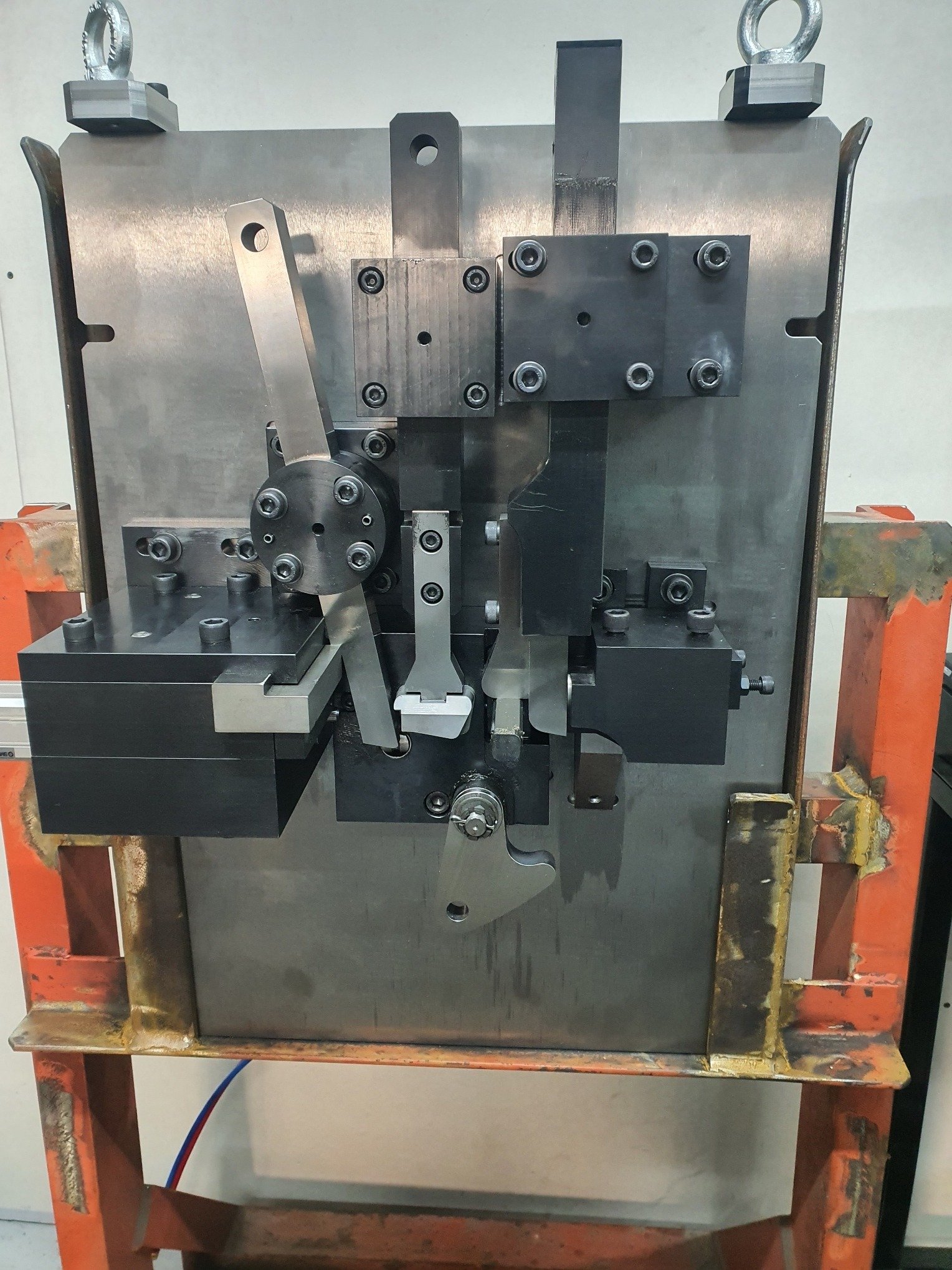 Metalowa forma z elementami mocującymi i ruchomymi, umieszczona na pomarańczowej ramie. Widoczne śruby, blachy i elementy konstrukcyjne.