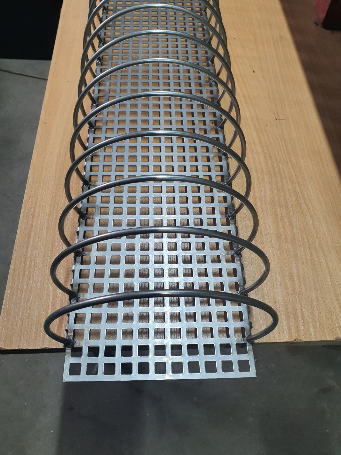 Metalowa konstrukcja z perforowanej blachy i spawanych okręgów, leżąca na drewnianej powierzchni. Widoczne ślady spawania.