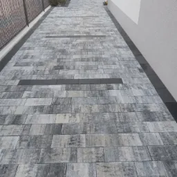 Ułożona ścieżka z prostokątnej kostki brukowej w odcieniach szarości, z ciemniejszymi wstawkami i obrzeżem, prowadząca wzdłuż ogrodzenia i ściany budynku.