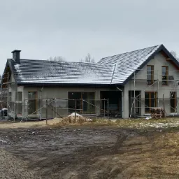 Surowy, nieotynkowany dom jednorodzinny w trakcie budowy, z rusztowaniami, czarną dachówką pokrytą śniegiem i błotnistym terenem wokół.