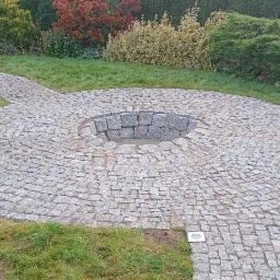 Okrągły placyk z kostki granitowej z centralnym zagłębieniem otoczonym kamieniami, połączony ścieżkami z kostki, wkomponowany w trawnik z ozdobnymi krzewami w tle.