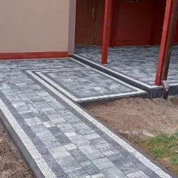 Nowa nawierzchnia z kostki brukowej w odcieniach szarości, z dekoracyjnymi obramowaniami, prowadząca do wejścia domu z czerwonymi słupami werandy, widoczny wózek budowlany i narzędzia ogrodowe.