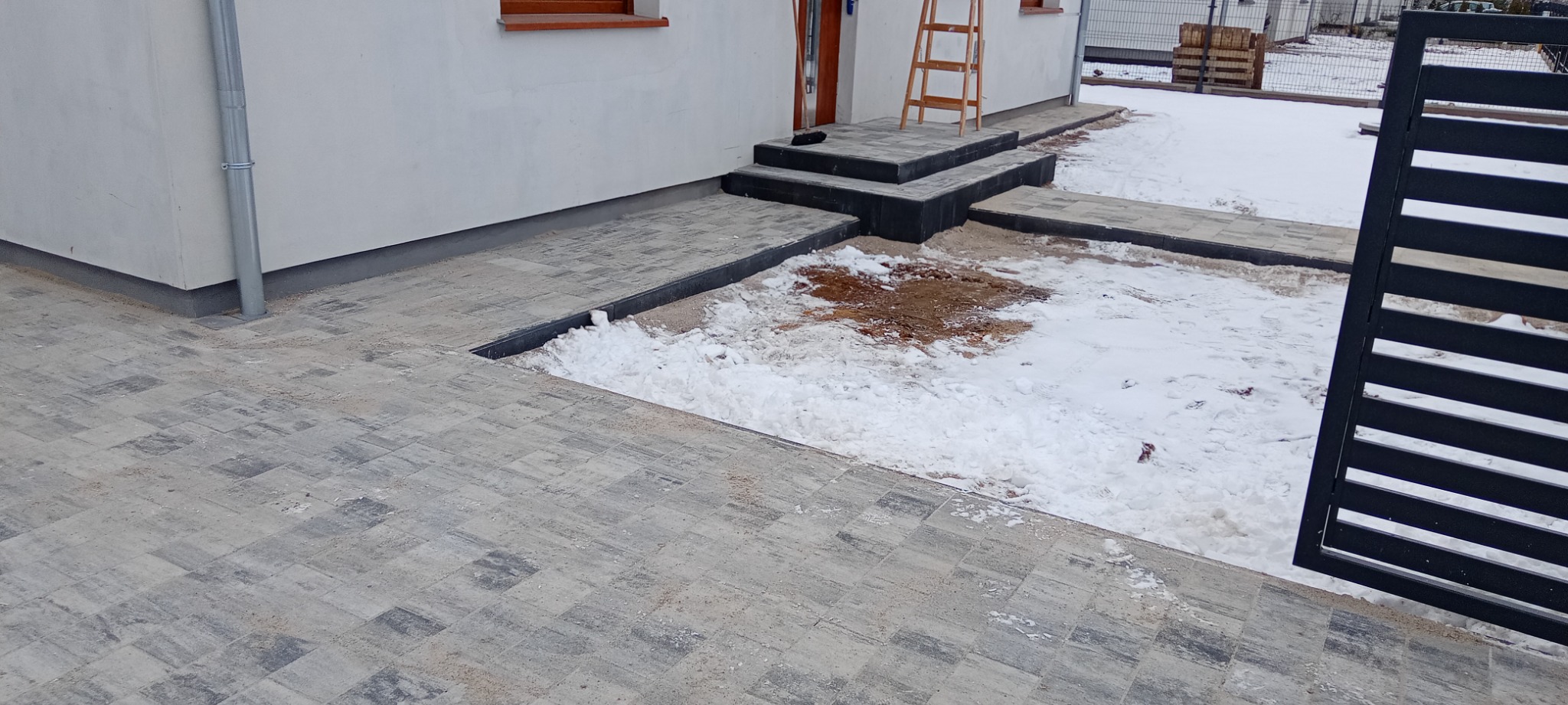 Ułożona kostka brukowa prowadząca do wejścia domu, częściowo pokryta śniegiem, widoczne schody i drabina oparta o ścianę.