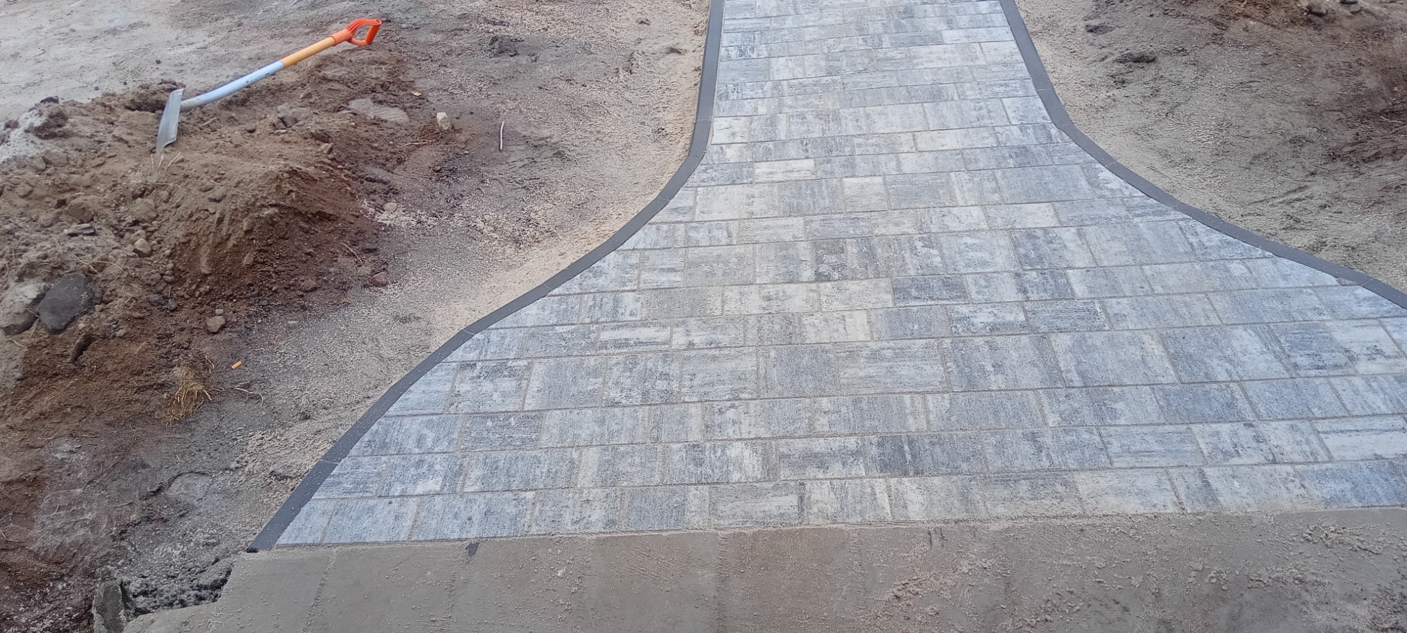 Świeżo ułożona ścieżka z szarej kostki brukowej w trakcie prac wykończeniowych, widoczna łopata w ziemi i piasek