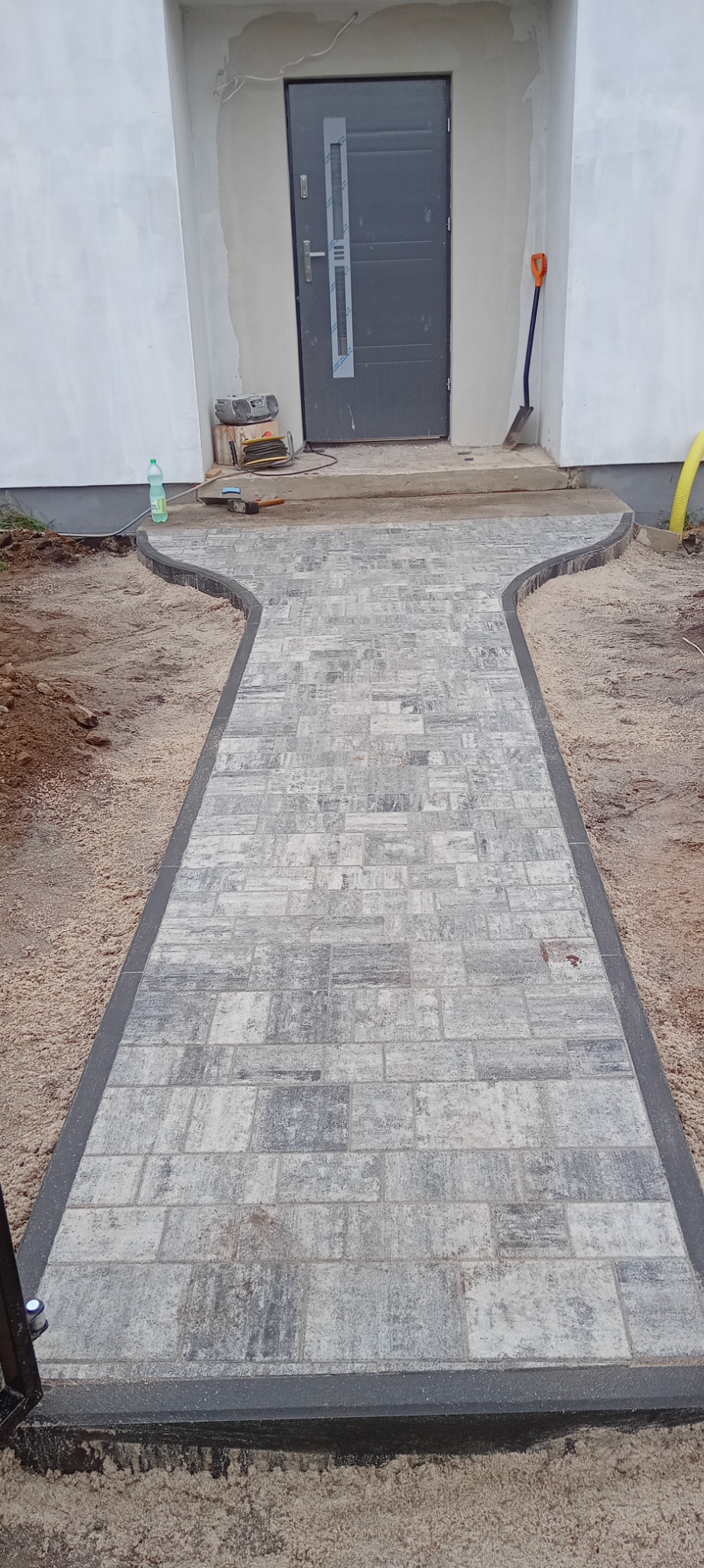 Ścieżka z szarej kostki brukowej prowadząca do drzwi wejściowych domu, obrzeża wykończone ciemnoszarą kostką, widoczne narzędzia i materiały budowlane w tle.