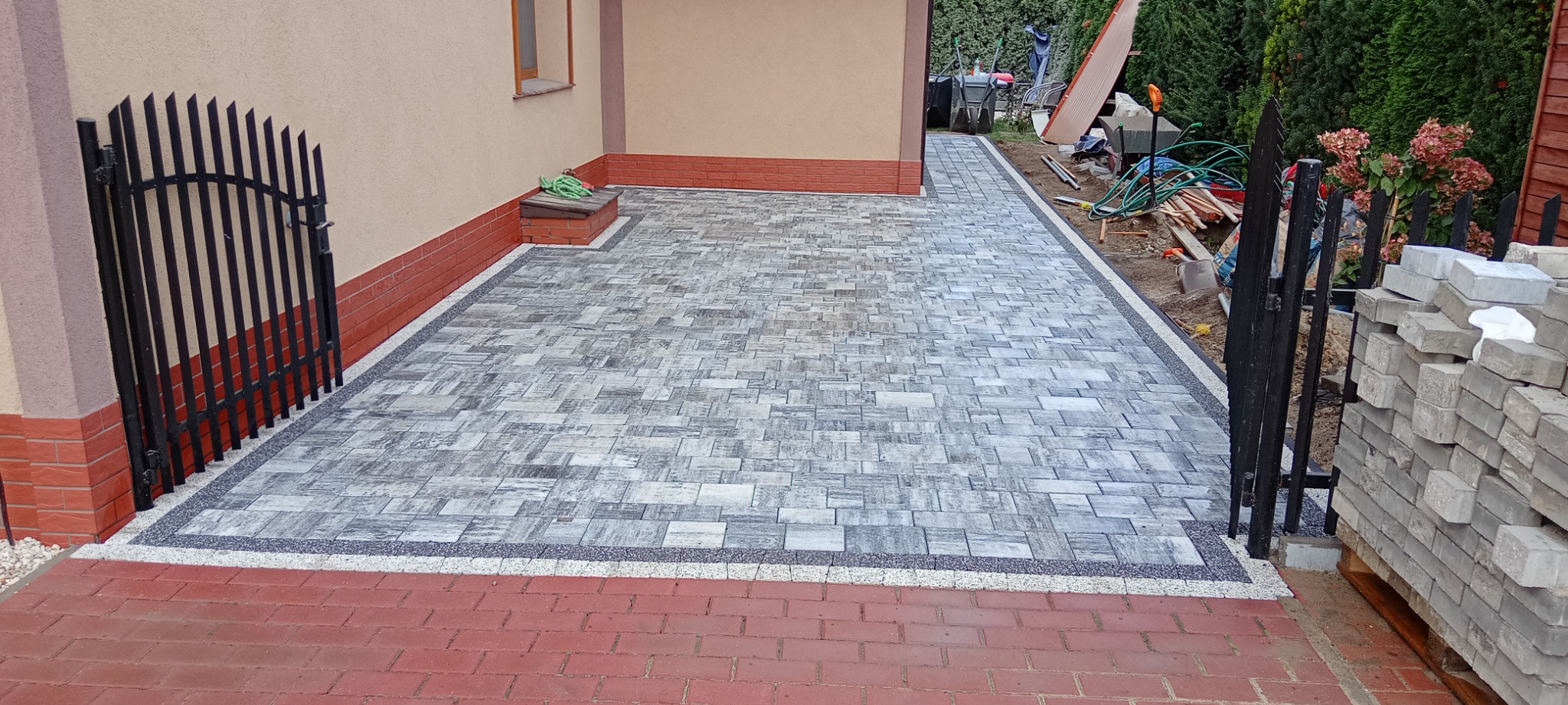Ułożona szara kostka brukowa na podwórku z dekoracyjnym obramowaniem z drobnego kamienia, widoczna brama z kutego żelaza, stos kostki brukowej i narzędzia w tle.