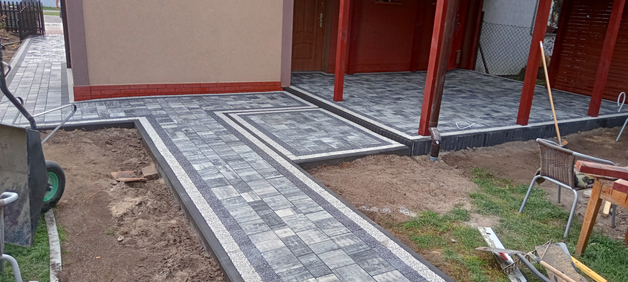 Nowa nawierzchnia z kostki brukowej w odcieniach szarości, z dekoracyjnymi obramowaniami, prowadząca do wejścia domu z czerwonymi słupami werandy, widoczny wózek budowlany i narzędzia ogrodowe.