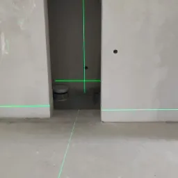 Wnętrze w stanie surowym z widocznymi laserowymi liniami poziomymi i pionowymi, wyznaczającymi płaszczyzny na ścianach i podłodze, z widocznym wiaderkiem farby w przejściu do innego pomieszczenia.