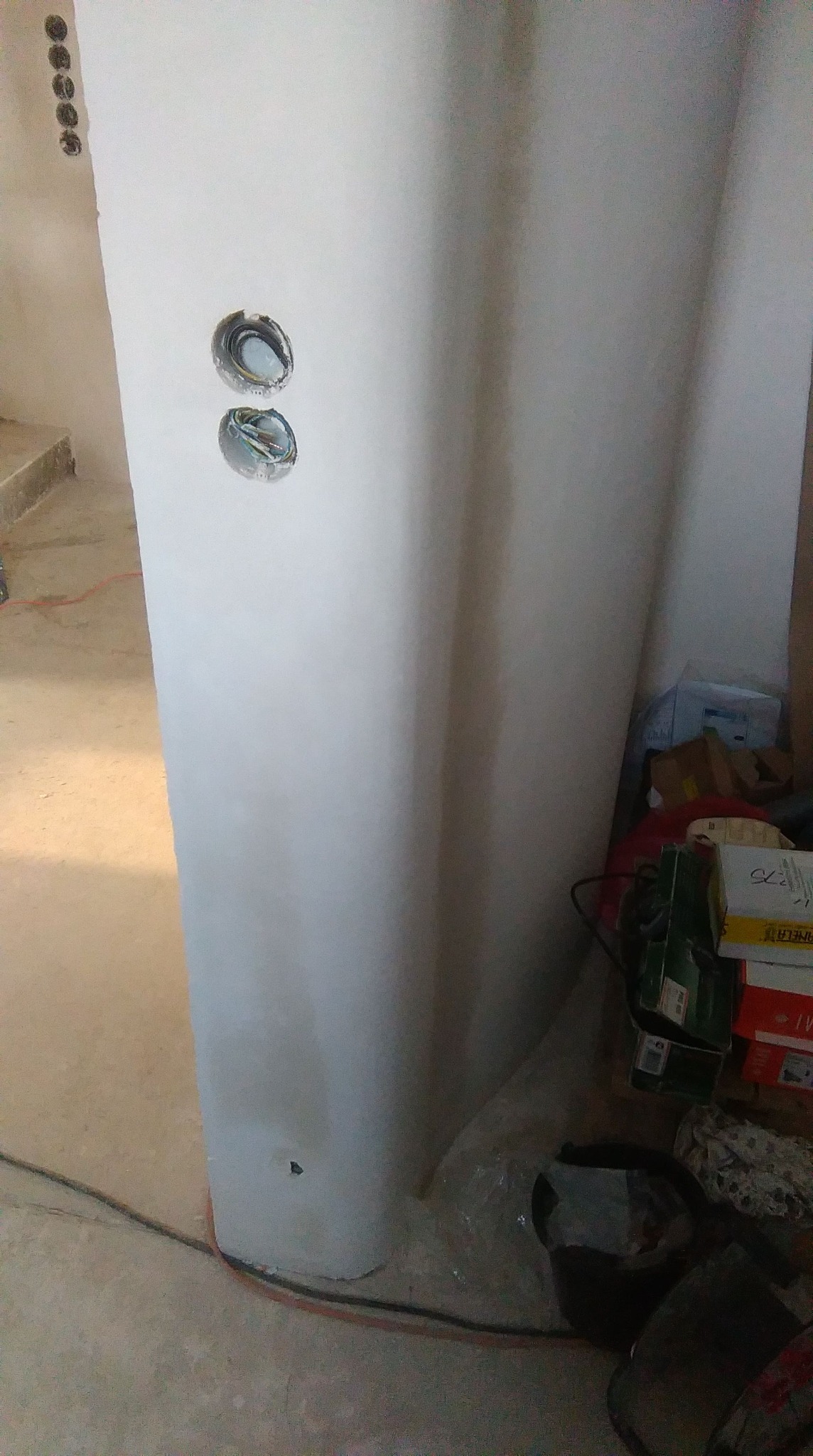 Świeżo otynkowana ściana w Kętach z widocznymi otworami pod instalację elektryczną, kable wystają ze ściany. W tle widoczne materiały budowlane.