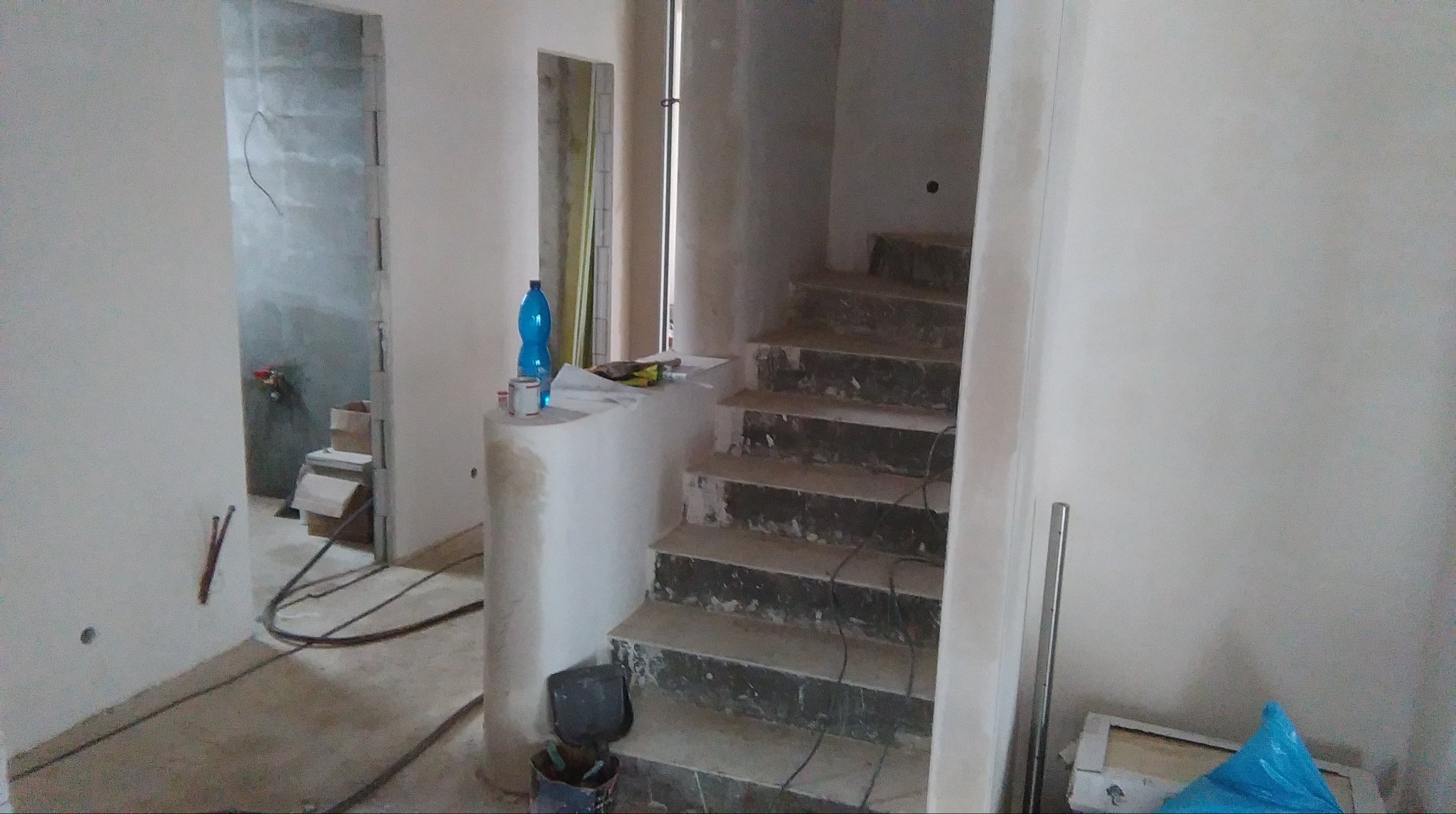 Wnętrze w trakcie prac wykończeniowych: świeżo otynkowane ściany, schody z widocznymi śladami remontu, butelka wody i puszki po farbie na półce.