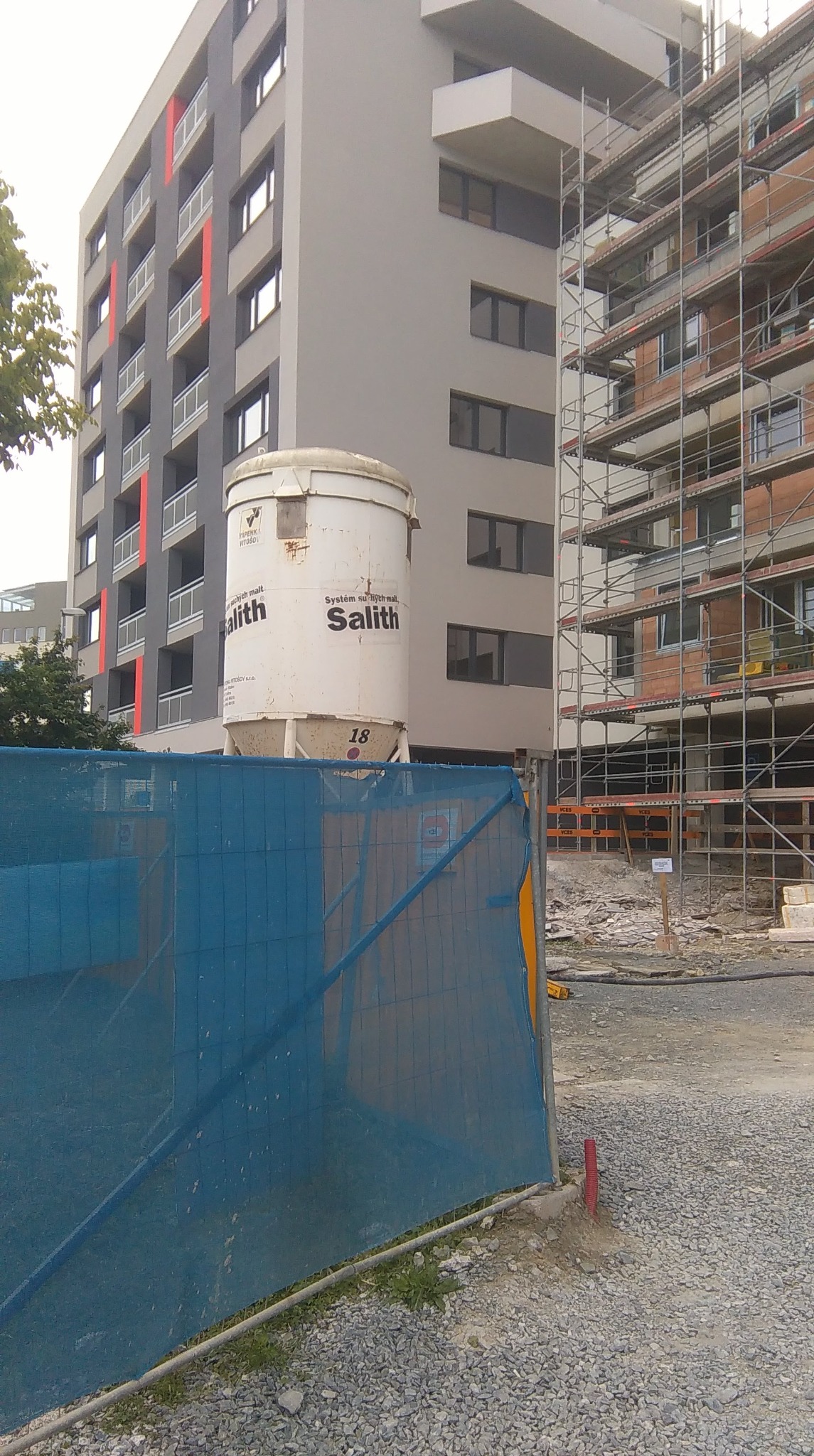 Silos na zaprawę firmy Salith na tle budynku w trakcie budowy z widocznymi rusztowaniami i niebieską siatką zabezpieczającą teren.