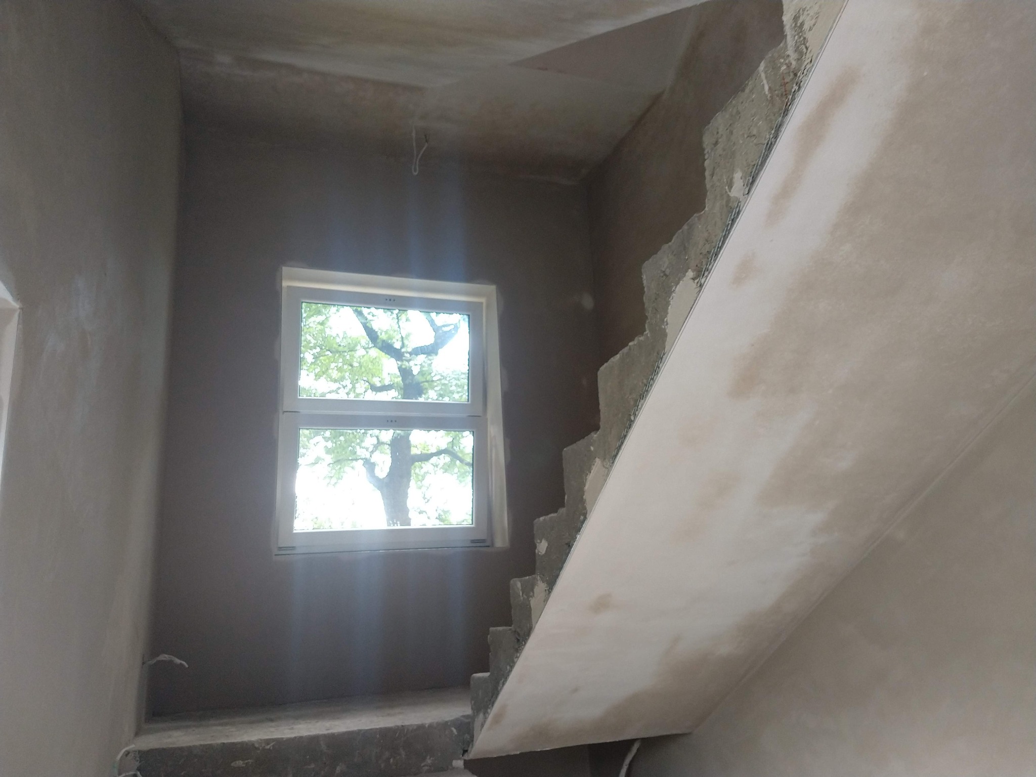 Wnętrze klatki schodowej w trakcie prac wykończeniowych: ściany pokryte świeżym tynkiem, widoczne surowe schody i okno z widokiem na drzewo.