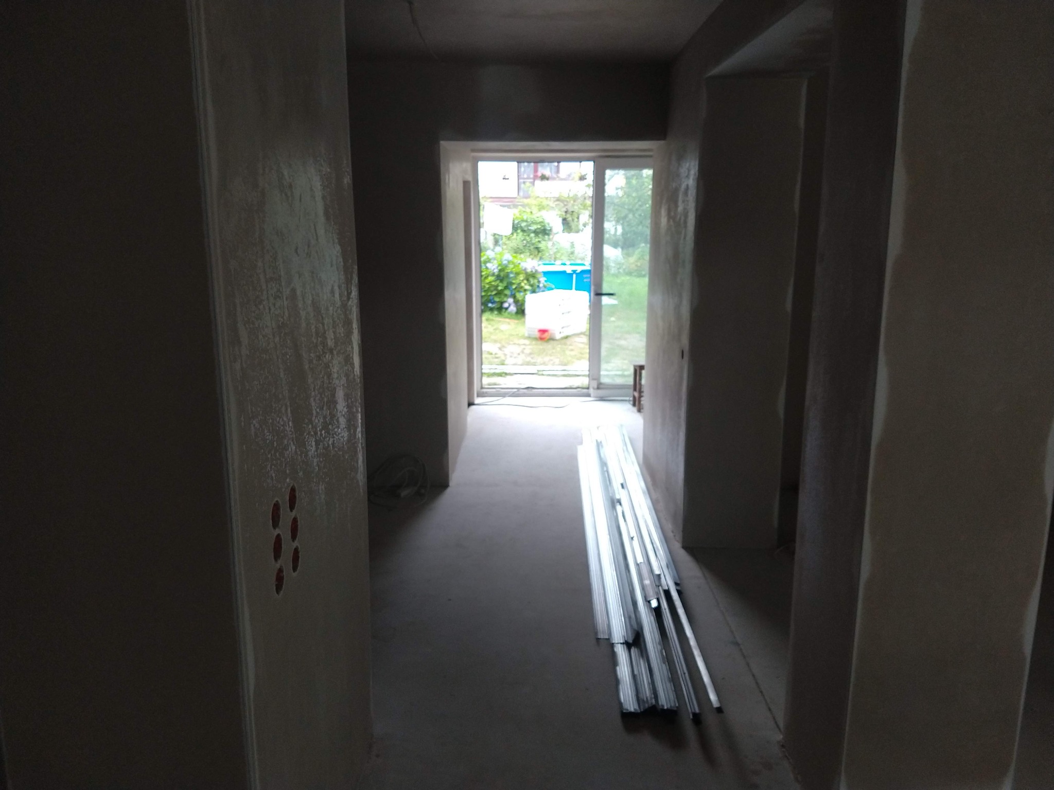 Korytarz w stanie surowym, ściany po nałożeniu tynku, widoczne puszki elektryczne, na podłodze leżą profile metalowe, w tle widok na ogród przez przeszklone drzwi.