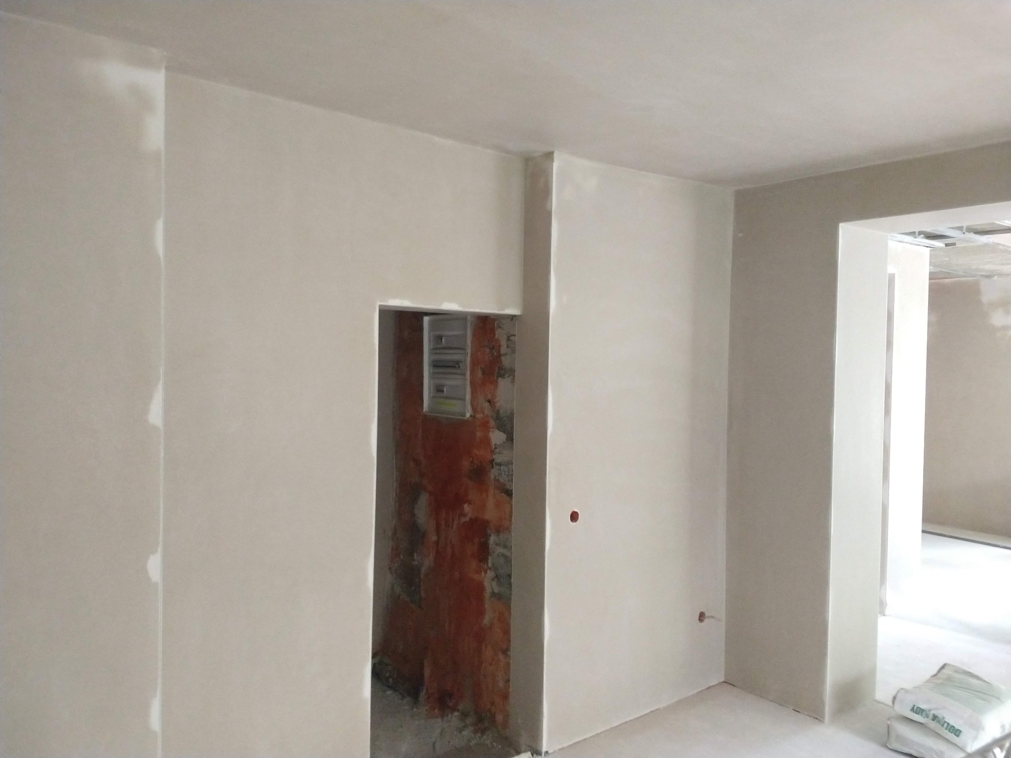 Świeżo położone, jasne tynki maszynowe na ścianach w surowym wnętrzu, widoczny otwór z odsłoniętą czerwoną cegłą i instalacją elektryczną oraz worki z zaprawą w rogu pomieszczenia.