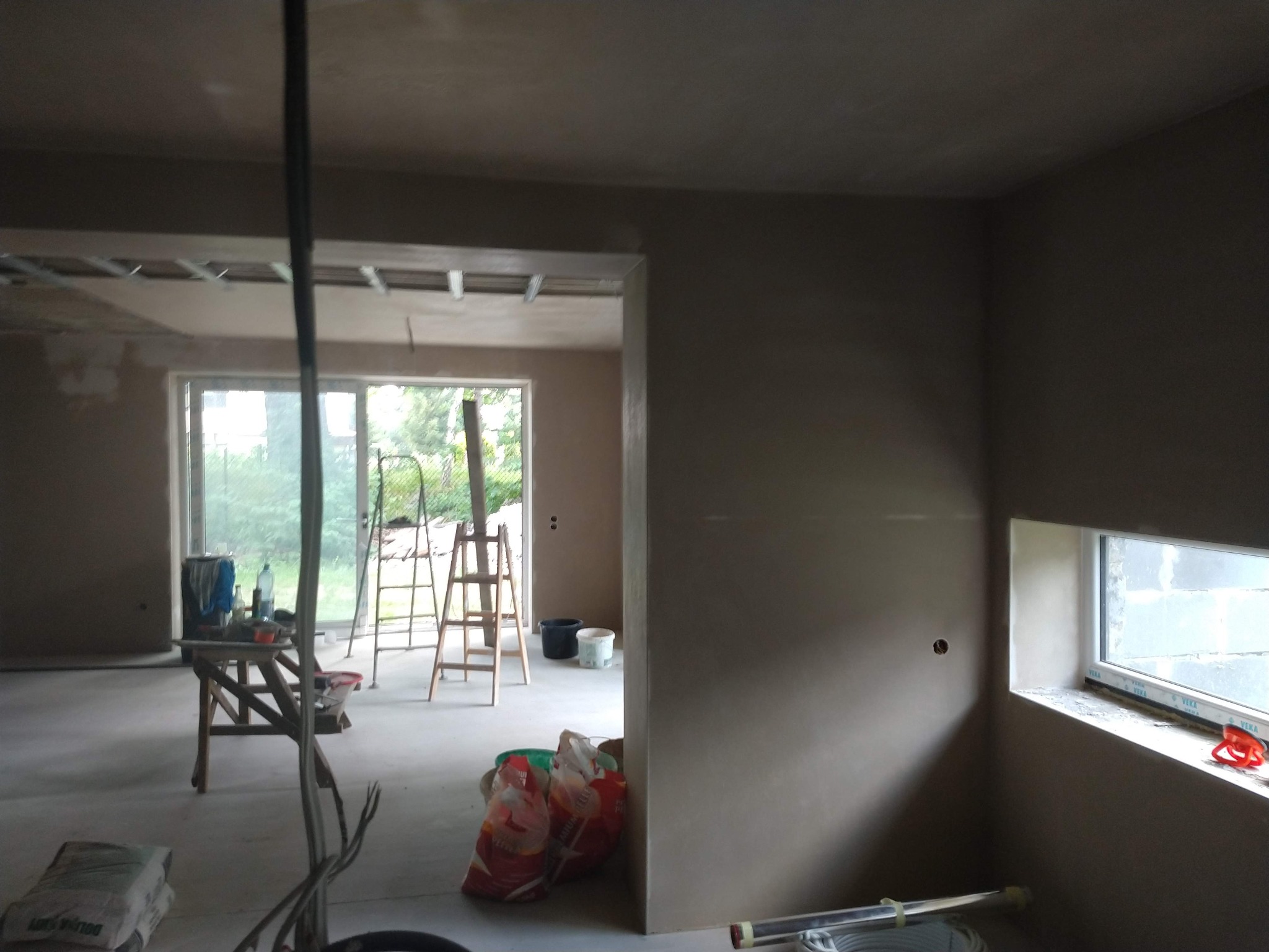 Wnętrze w trakcie prac wykończeniowych: świeżo nałożone tynki na ścianach, widoczne worki z zaprawą, drabina i prowizoryczny stół roboczy przy oknie, częściowo widoczne instalacje pod sufitem.