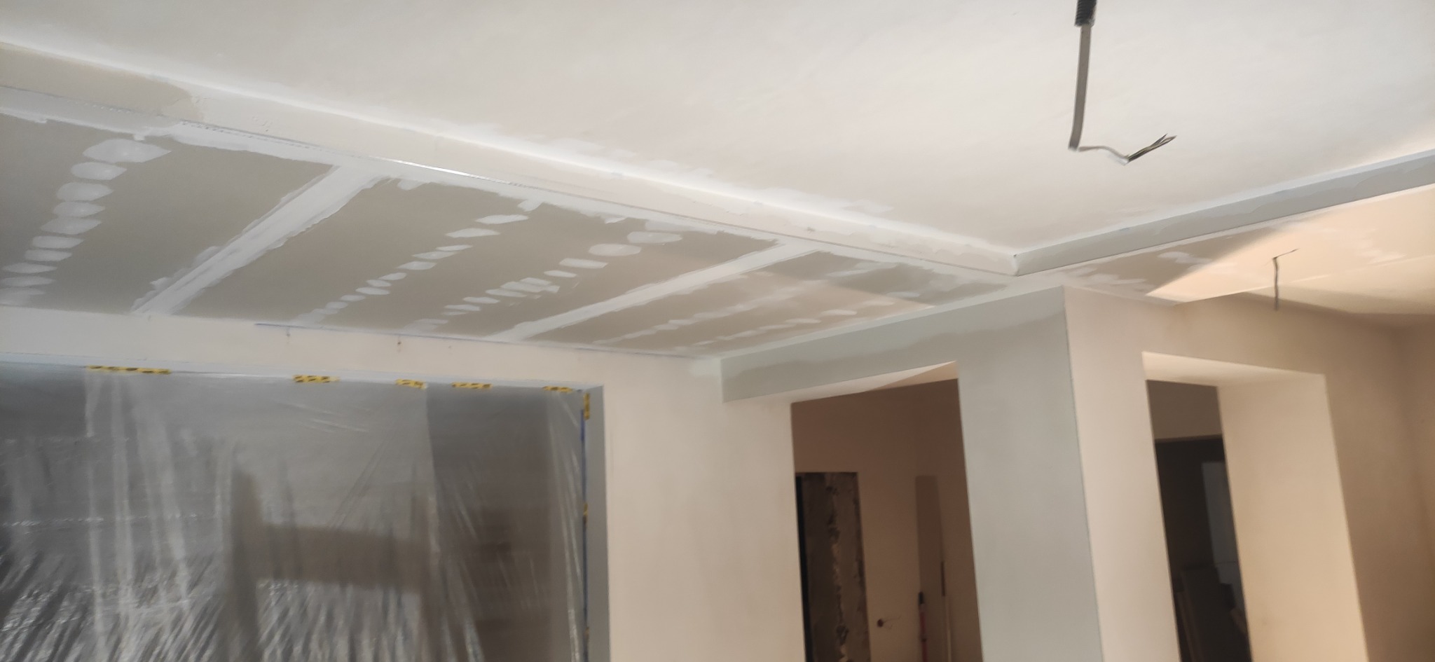 Sufit z płyt gipsowo-kartonowych w trakcie szpachlowania łączeń, widoczne fragmenty instalacji elektrycznej wystające z sufitu, pomieszczenie w stanie surowym z widoczną folią ochronną na ścianie.