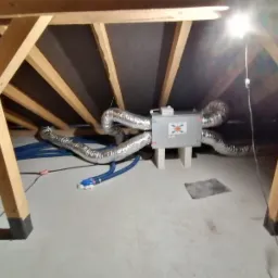 Szare urządzenie rekuperacyjne z podłączonymi elastycznymi przewodami wentylacyjnymi w surowym pomieszczeniu na poddaszu z widoczną konstrukcją drewnianą dachu i niebieskimi rurami.