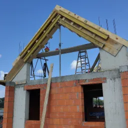 Budowa domu w stanie surowym otwartym: ściany z cegły, betonowe słupy, drewniana konstrukcja dachu z deskami, drabina, niebieskie niebo z chmurami w tle.