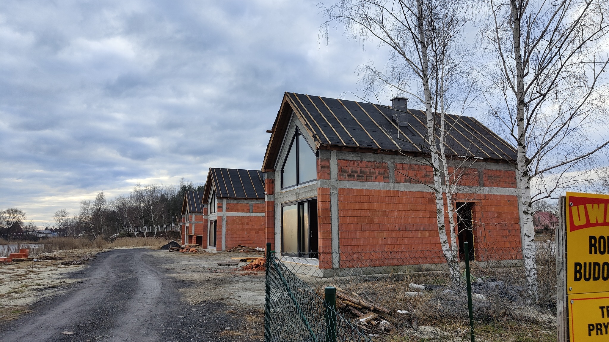 Surowe domy z czerwonej cegły z czarnym dachem w trakcie budowy, widoczne przez metalowe ogrodzenie. Drzewa i pochmurne niebo w tle.