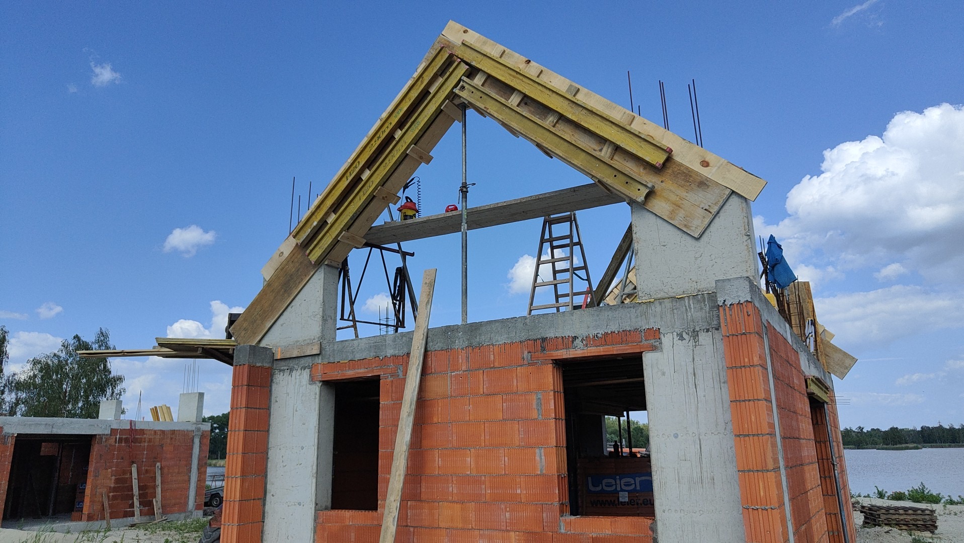 Budowa domu w stanie surowym otwartym: ściany z cegły, betonowe słupy, drewniana konstrukcja dachu z deskami, drabina, niebieskie niebo z chmurami w tle.