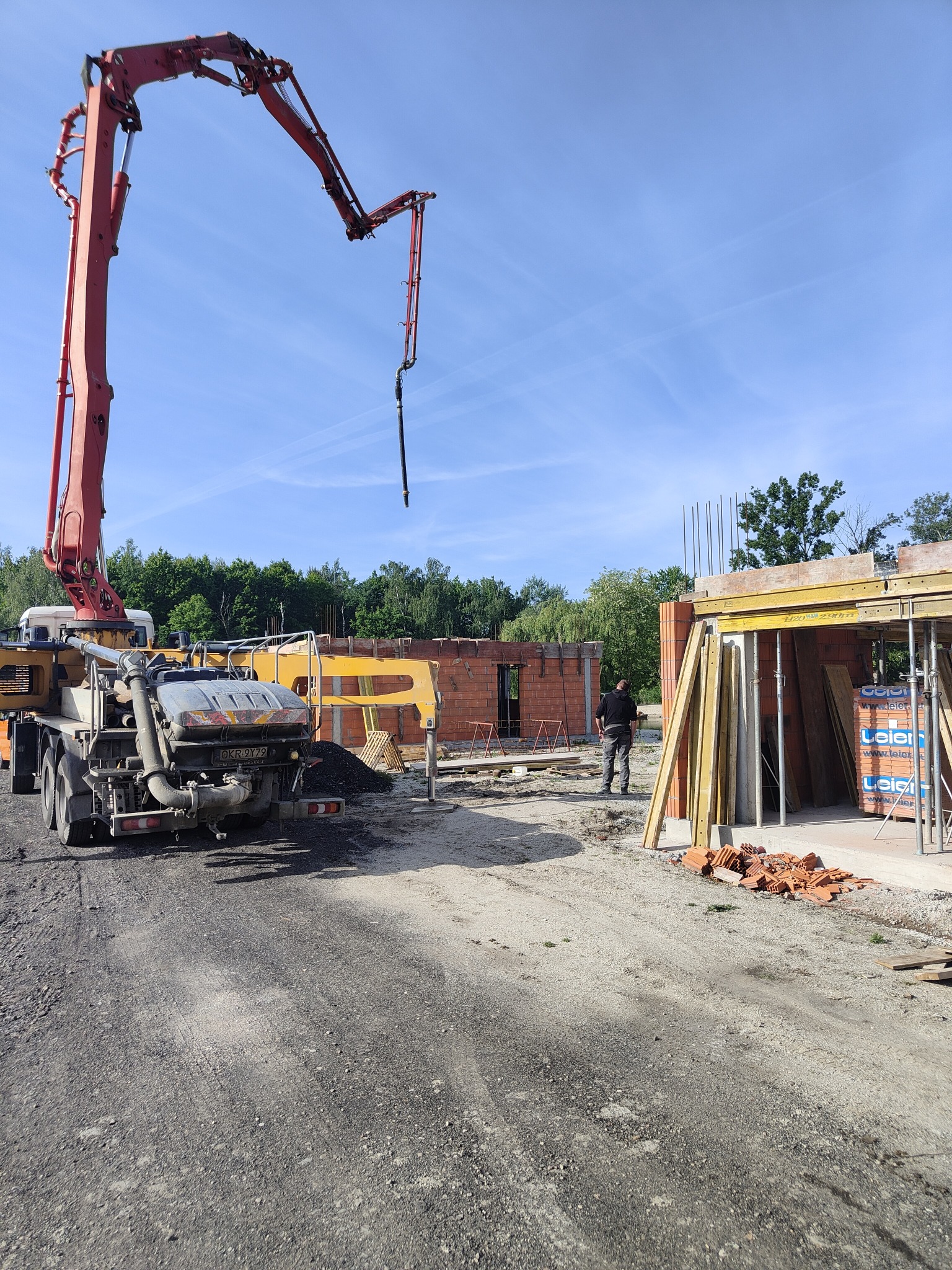 Budowa domu w trakcie: betonowa pompa podaje mieszankę do wznoszonych ścian z czerwonej cegły. Widoczny pracownik, szalunki i materiały budowlane.