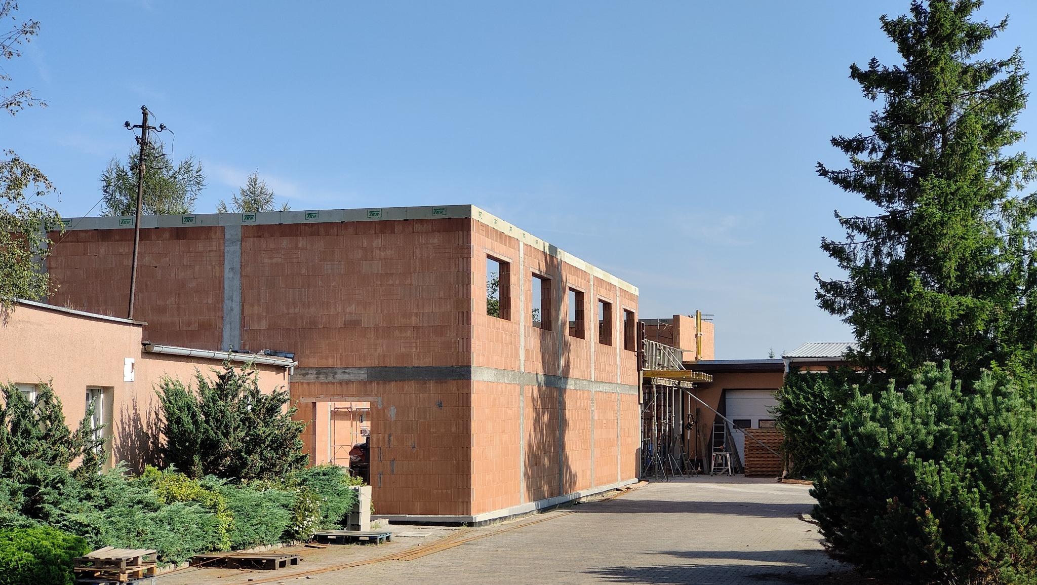 Budowa domu z pustaków ceramicznych, widoczne otwory okienne i przygotowane szalunki. Budynek w stanie surowym otwartym, otoczony zielenią i innymi budynkami.