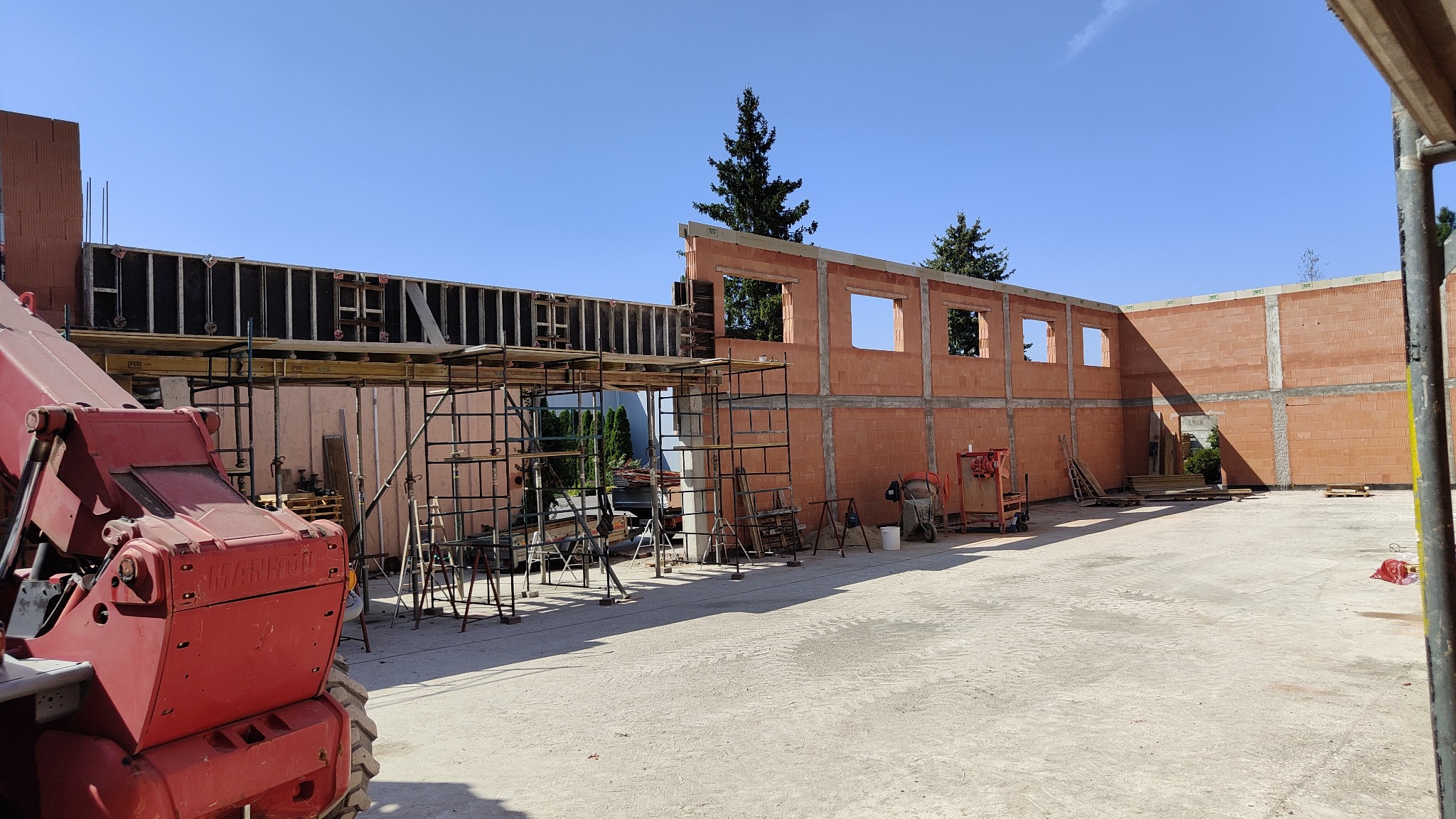 Budowa domu z czerwonej cegły, widoczne rusztowania i szalunki. Częściowo wzniesione ściany z otworami okiennymi. Na pierwszym planie czerwony podnośnik.