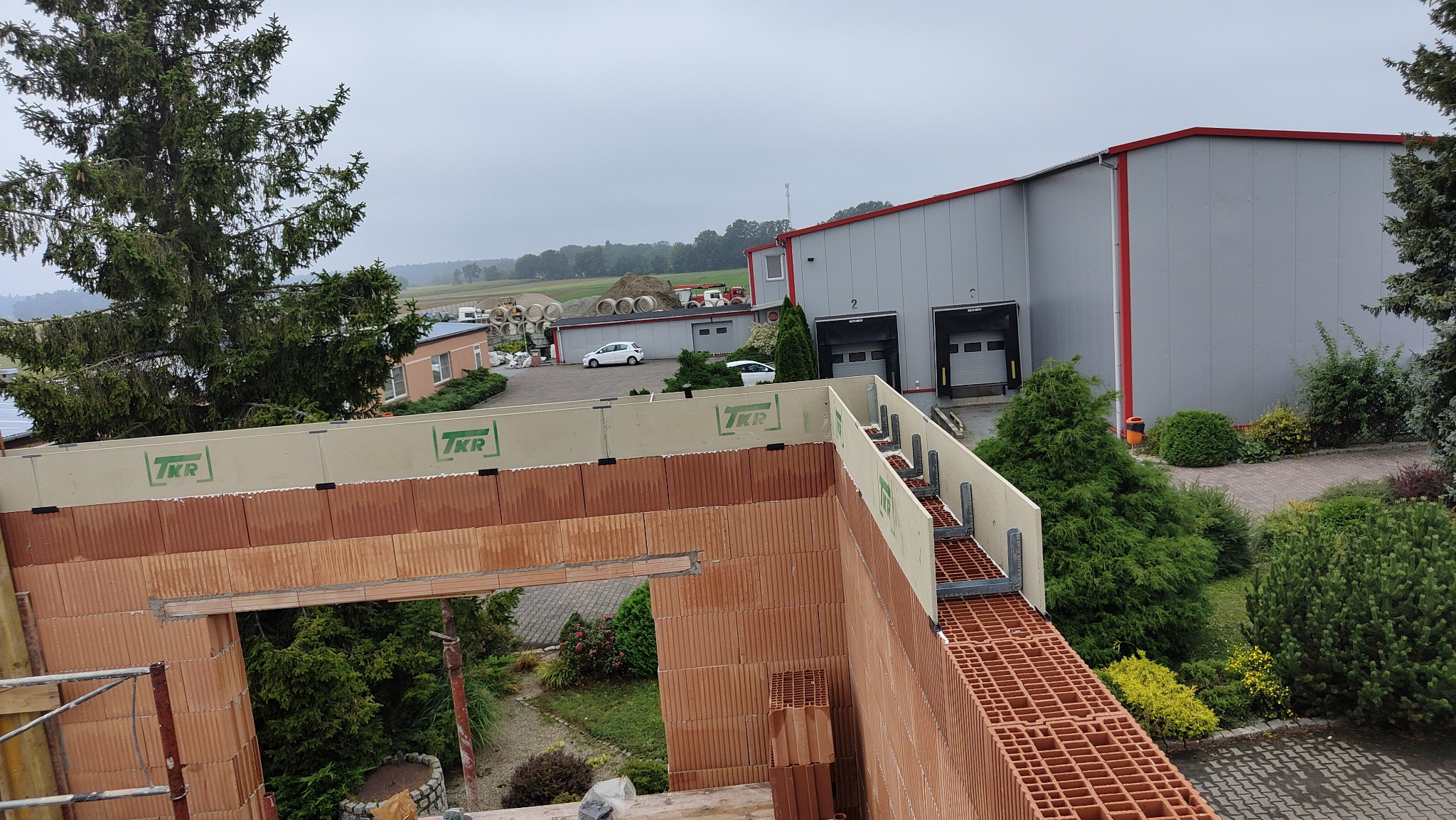 Wznoszone ściany budynku z pustaków ceramicznych, widoczne elementy szalunku i zbrojenia. W tle zabudowania gospodarcze i zieleń.