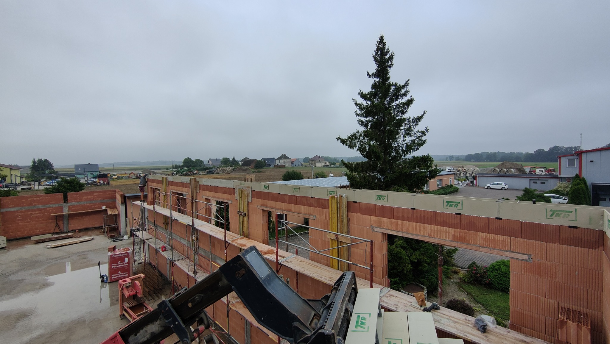 Wznoszenie ścian domu z pustaków ceramicznych Porotherm na tle krajobrazu. Widoczne rusztowania, elementy konstrukcyjne i sprzęt budowlany. Pochmurne niebo.