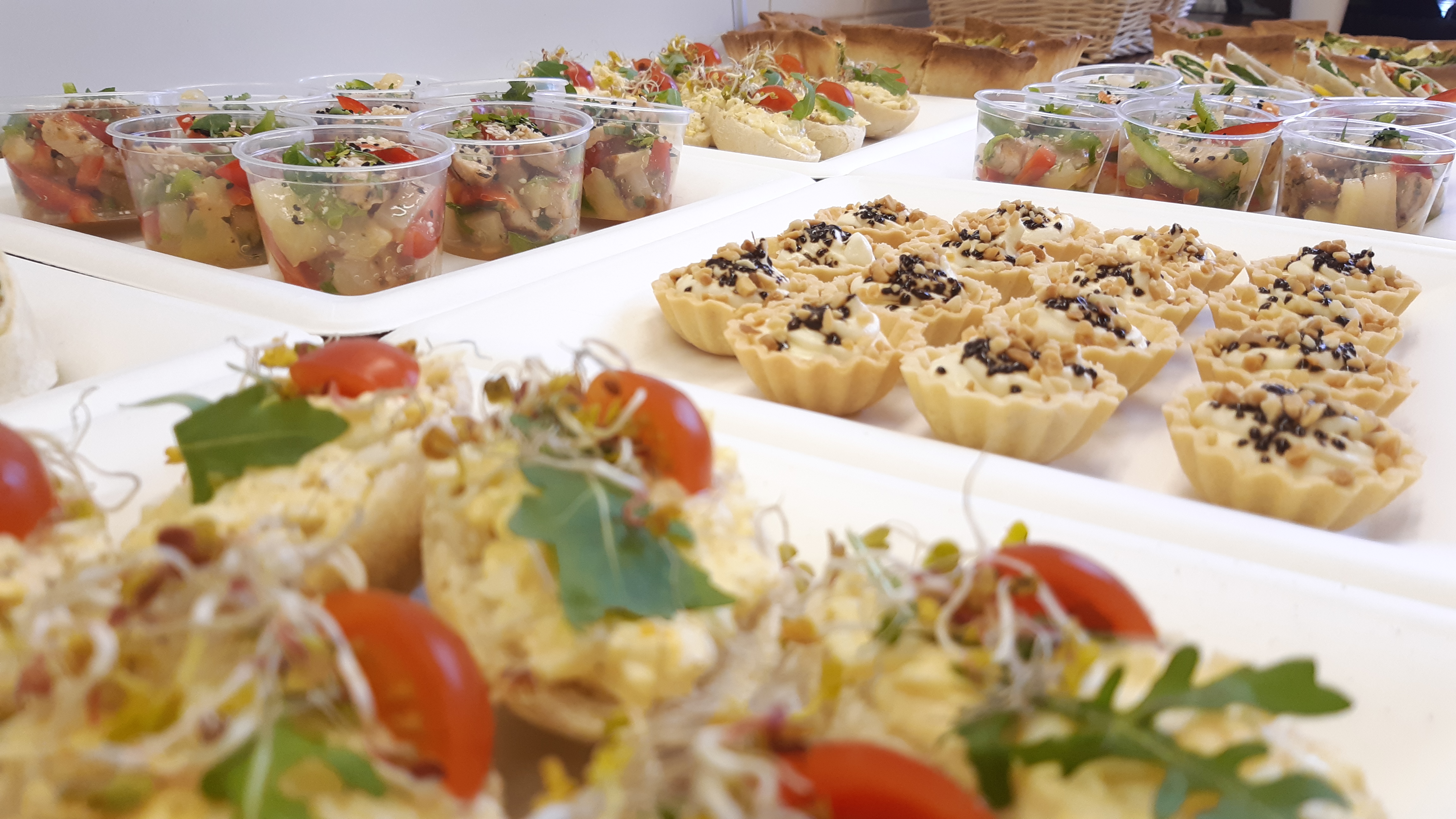 Różnorodny wybór przekąsek cateringowych: sałatki w kubeczkach, tartaletki z kremem i orzechami, kanapki z pastą jajeczną, pomidorkami i kiełkami na białych tacach.