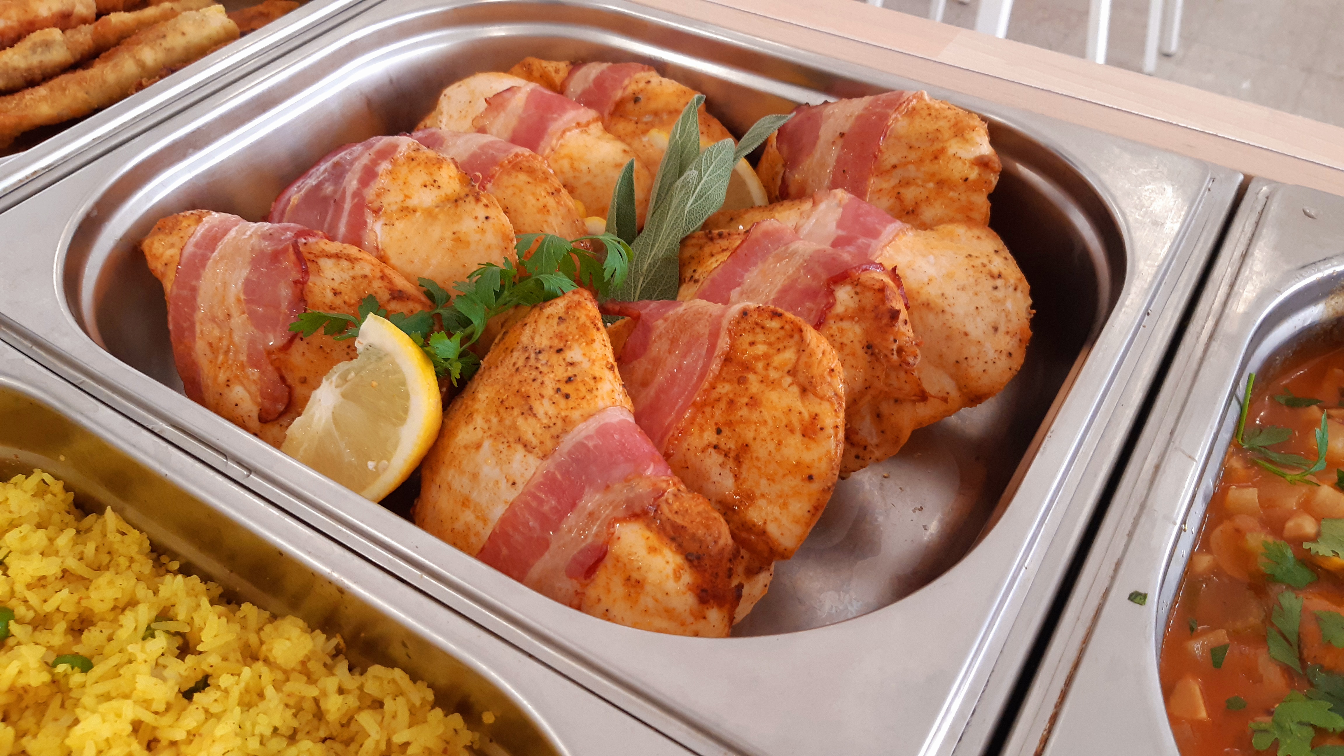 Catering w Warszawie: Metalowa taca z pieczonymi roladkami z kurczaka owiniętymi boczkiem, udekorowanymi cytryną, pietruszką i liśćmi szałwii, obok ryż i gulasz.