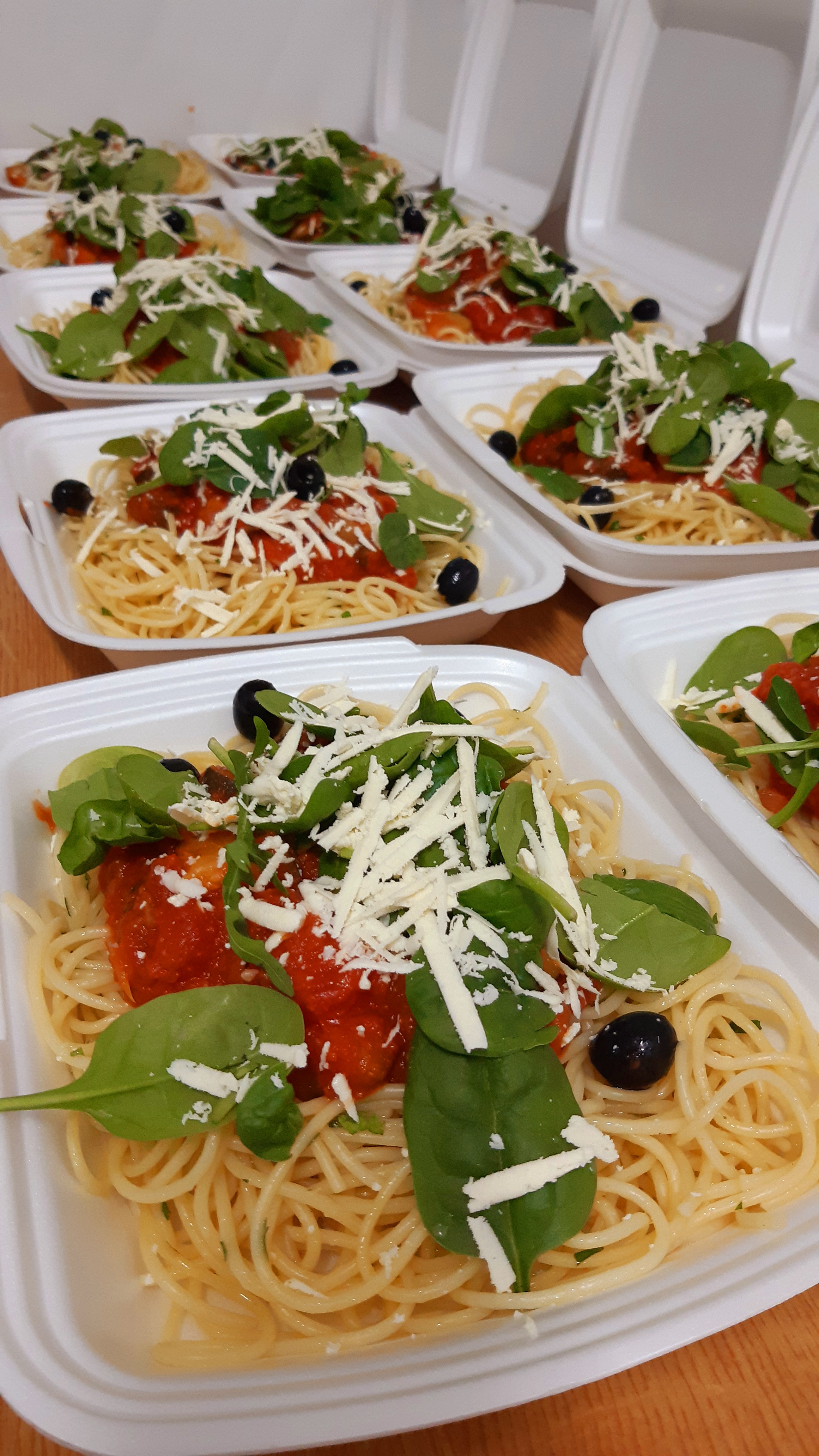 Spaghetti z sosem pomidorowym, szpinakiem, serem i oliwkami w białych pojemnikach cateringowych na drewnianym stole, gotowe do wydania.