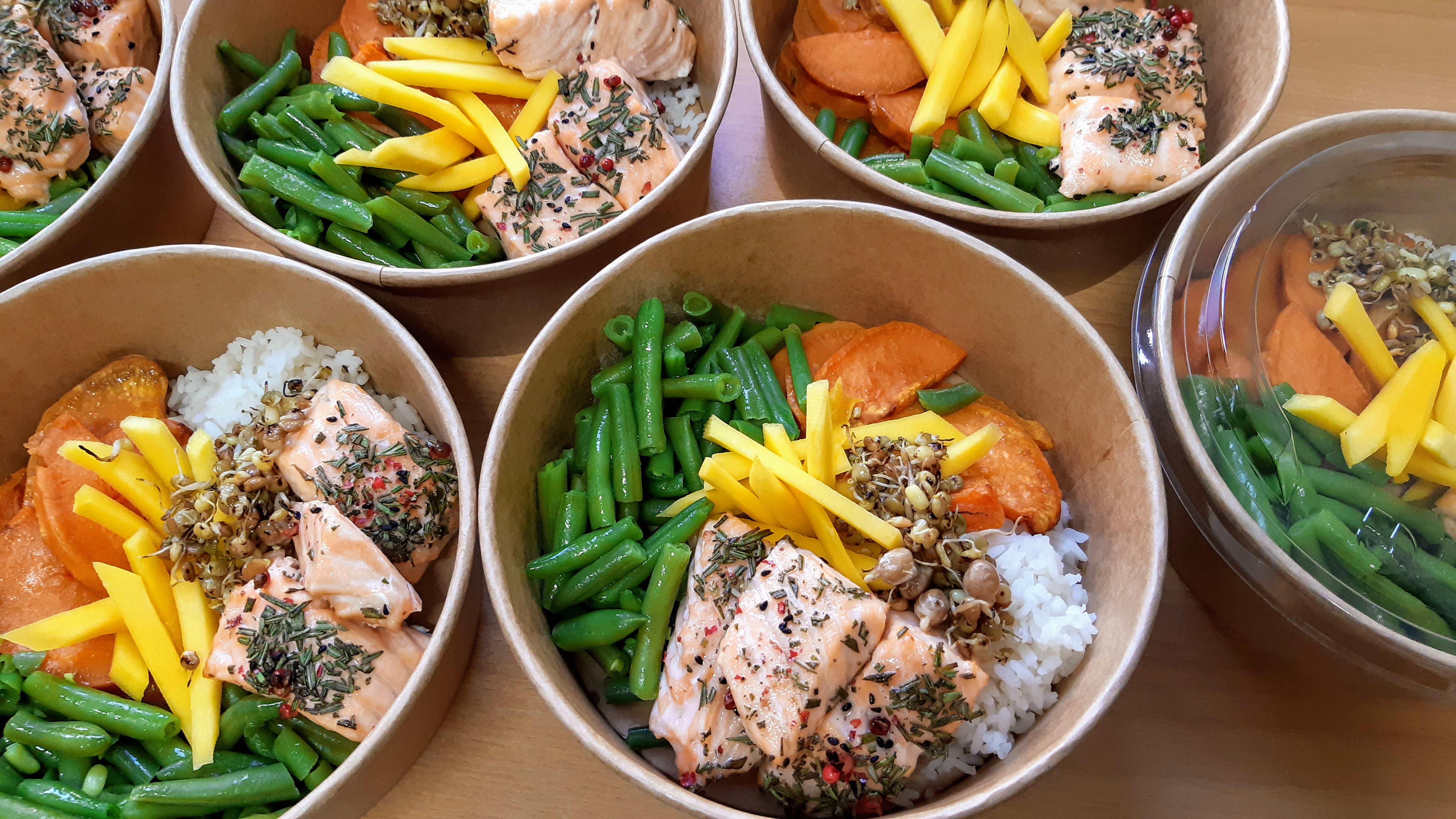 Lunchboxy z grillowanym łososiem posypanym ziołami, fasolką szparagową, mango i kiełkami na ryżu lub batatach, w papierowych miskach.