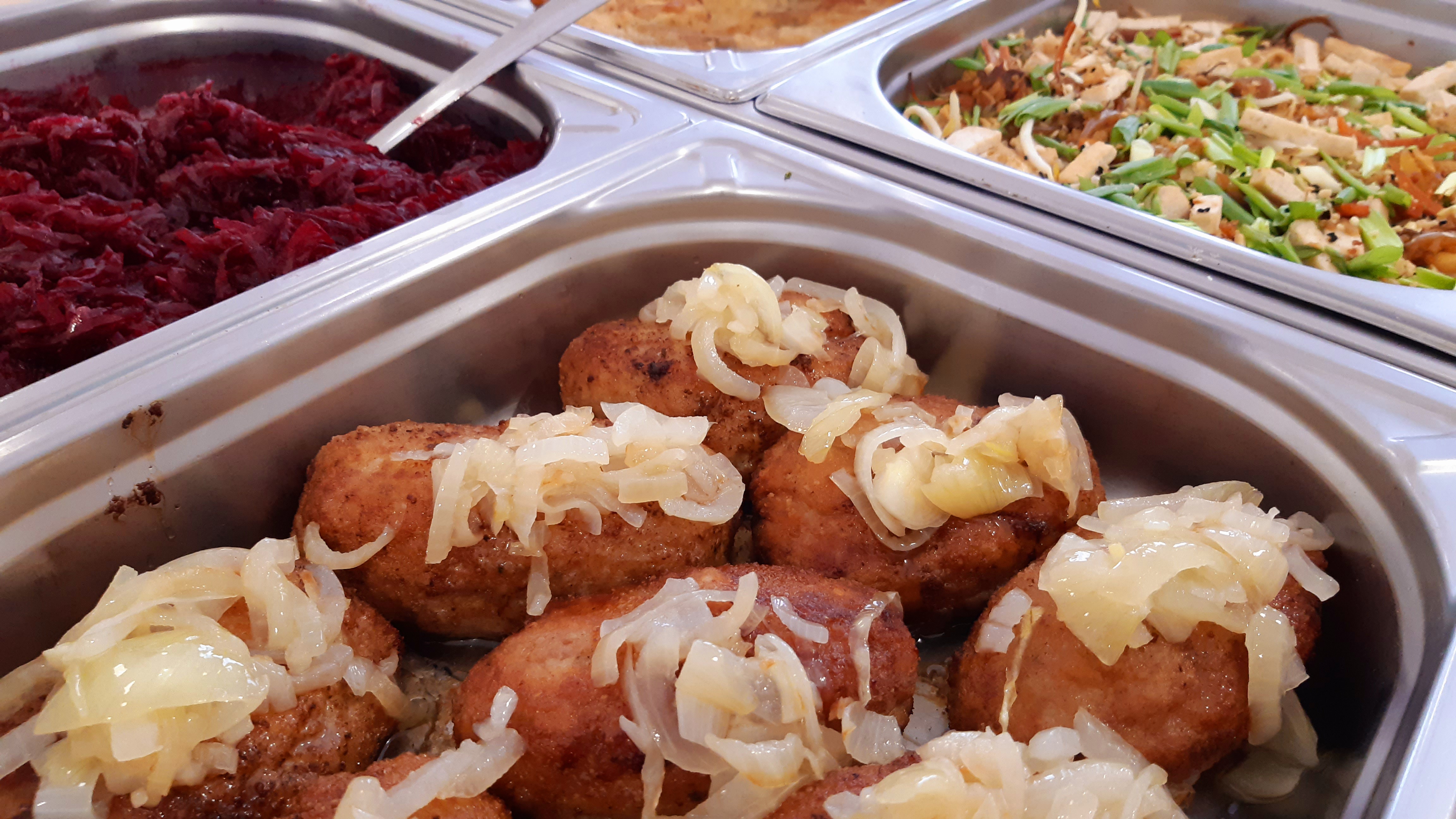 Metalowy pojemnik cateringowy z klopsikami polanymi duszoną cebulą, obok surówka z czerwonej kapusty i sałatka azjatycka.